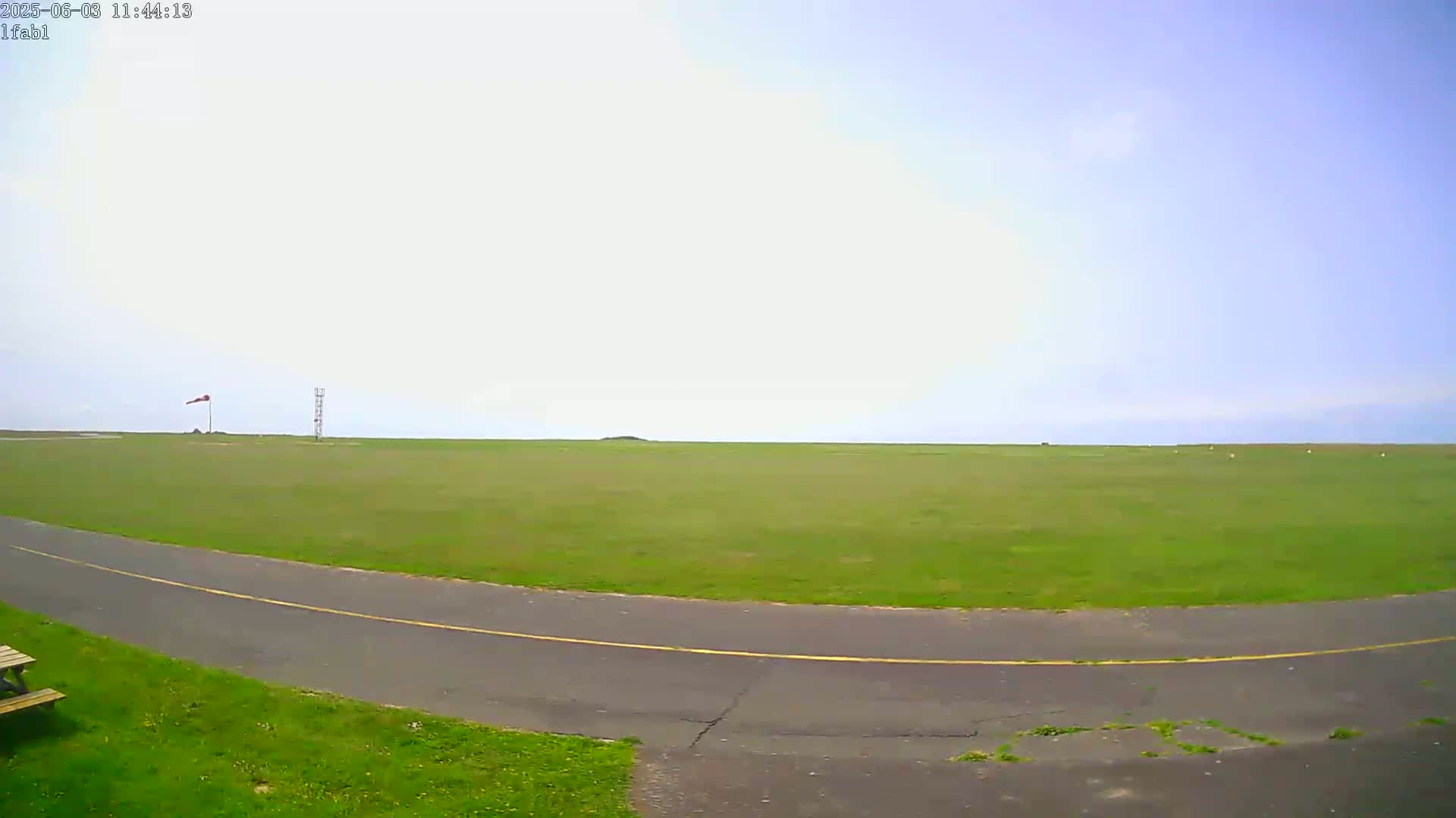 A mostly clear sky overlooks a grassy airfield with a paved road curving into the foreground.