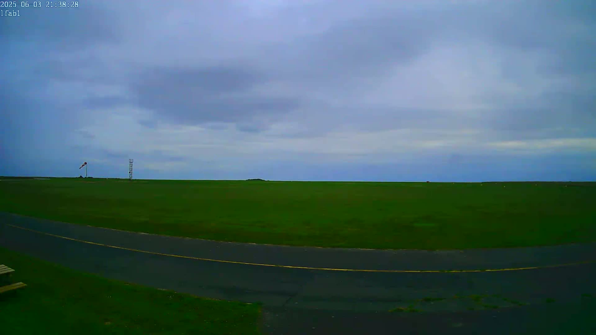 A vast green field stretches to a horizon under a mostly cloudy sky, with a paved road curving through the foreground.