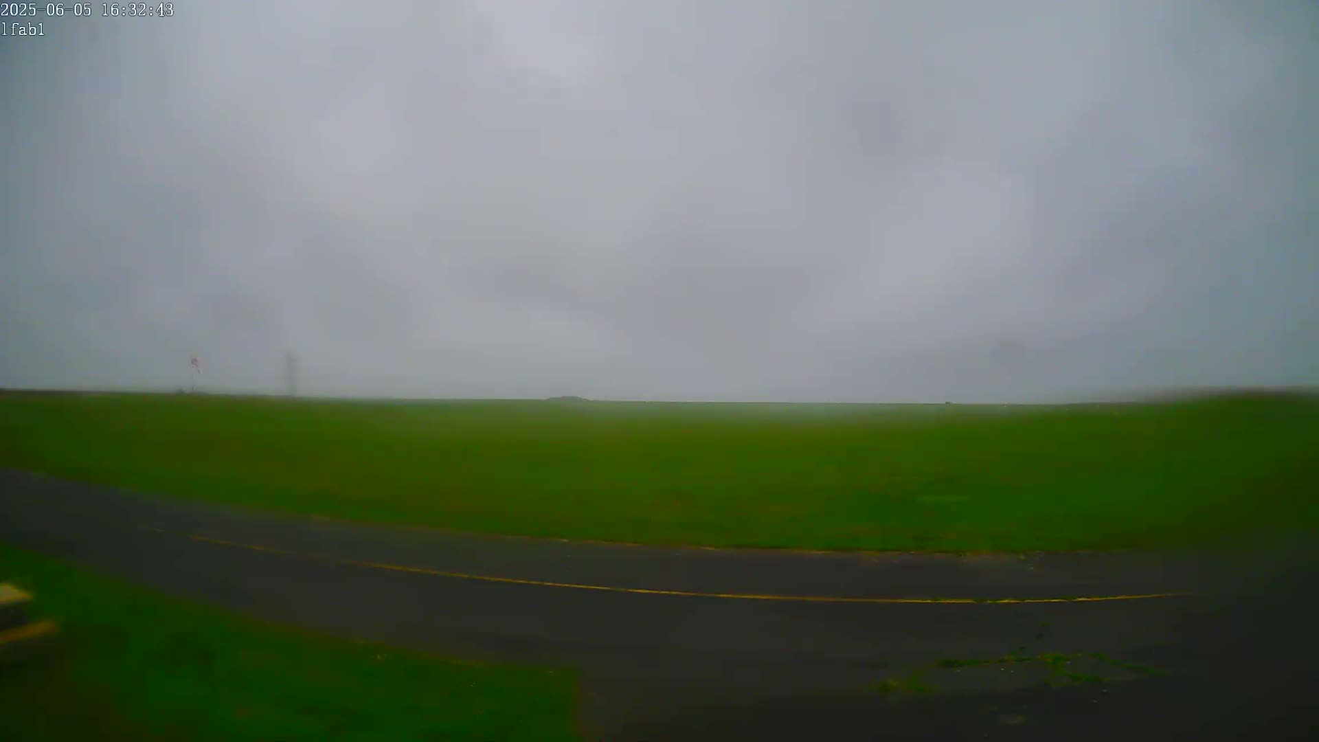 A hazy, overcast sky hangs over a flat expanse of green grass and a dark asphalt road.