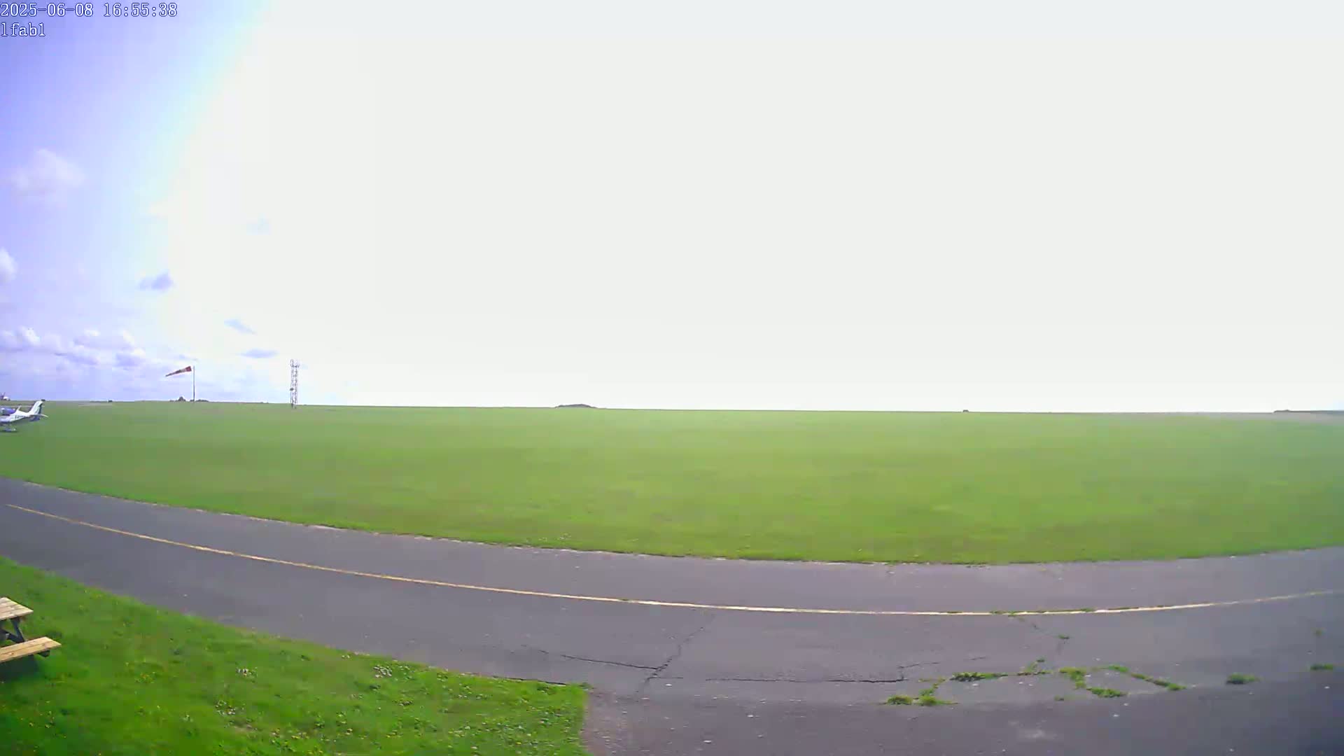 A small airplane is parked on a grassy airfield under a mostly cloudy sky, with a paved taxiway curving into view.