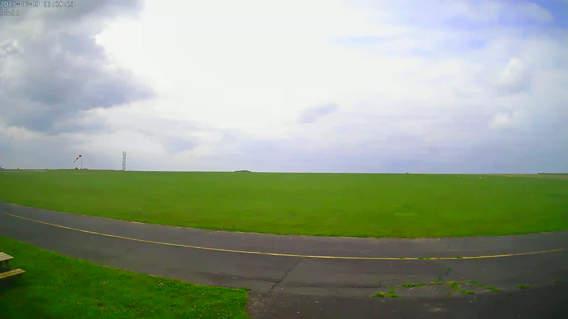 A grassy airfield under a partly cloudy sky features a paved road curving through the field, and a windsock and tower are visible in the distance.