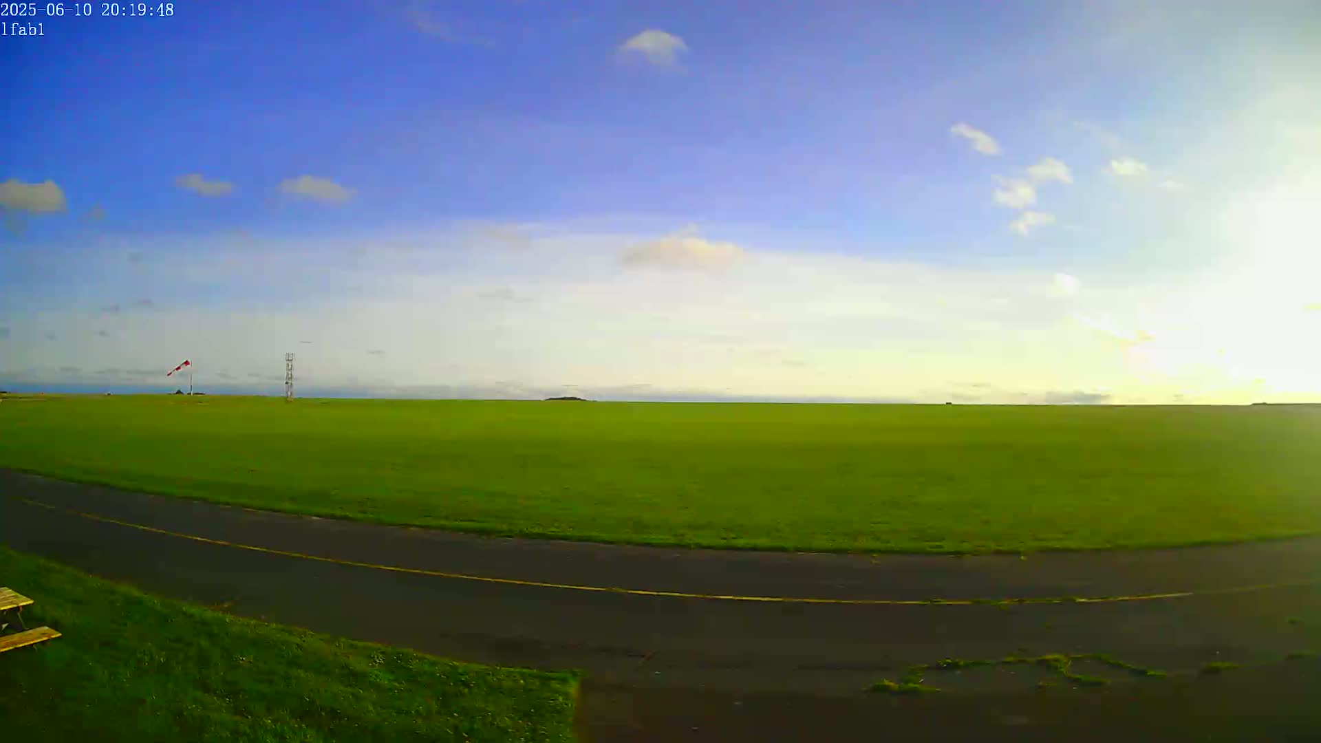 A vast green field stretches to the horizon under a mostly clear, blue sky with a few scattered clouds, and a paved road curves through the bottom of the image.