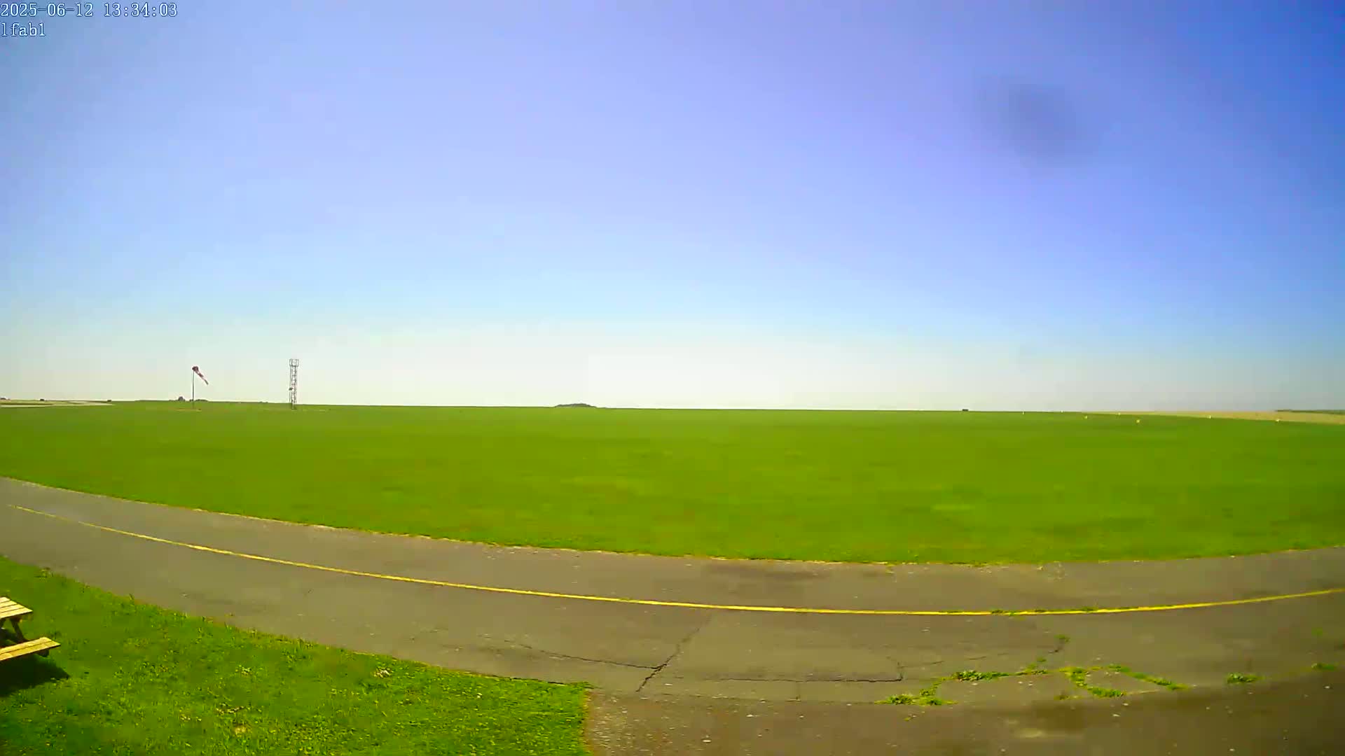 A vast green field, under a clear blue sky, is bordered by a paved path with a yellow line.