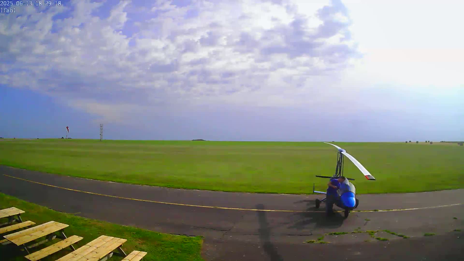 A person is working on a blue autogyro on a grassy airfield under a partly cloudy sky.