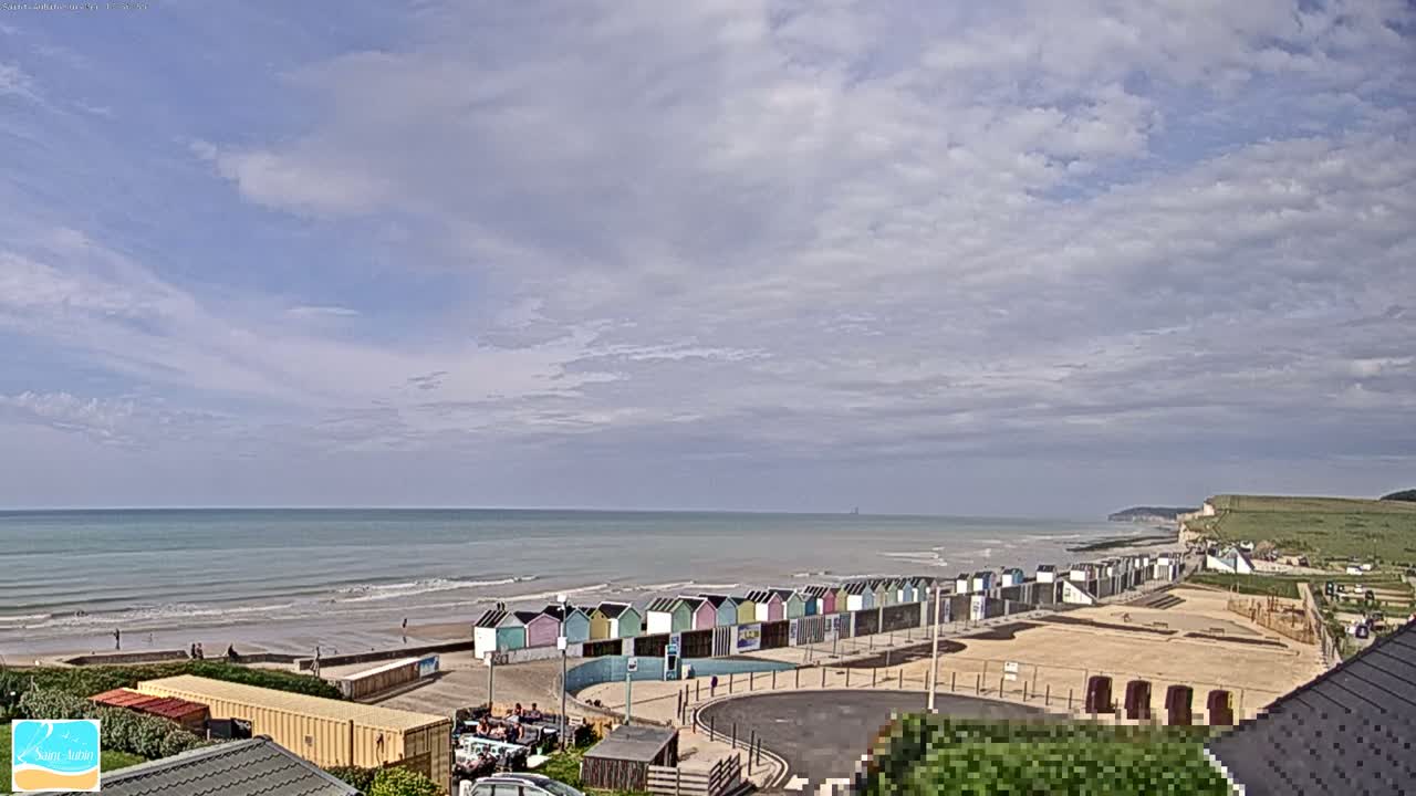 A beach with colorful beach huts is visible under a partly cloudy sky, with calm ocean waves rolling onto the shore.