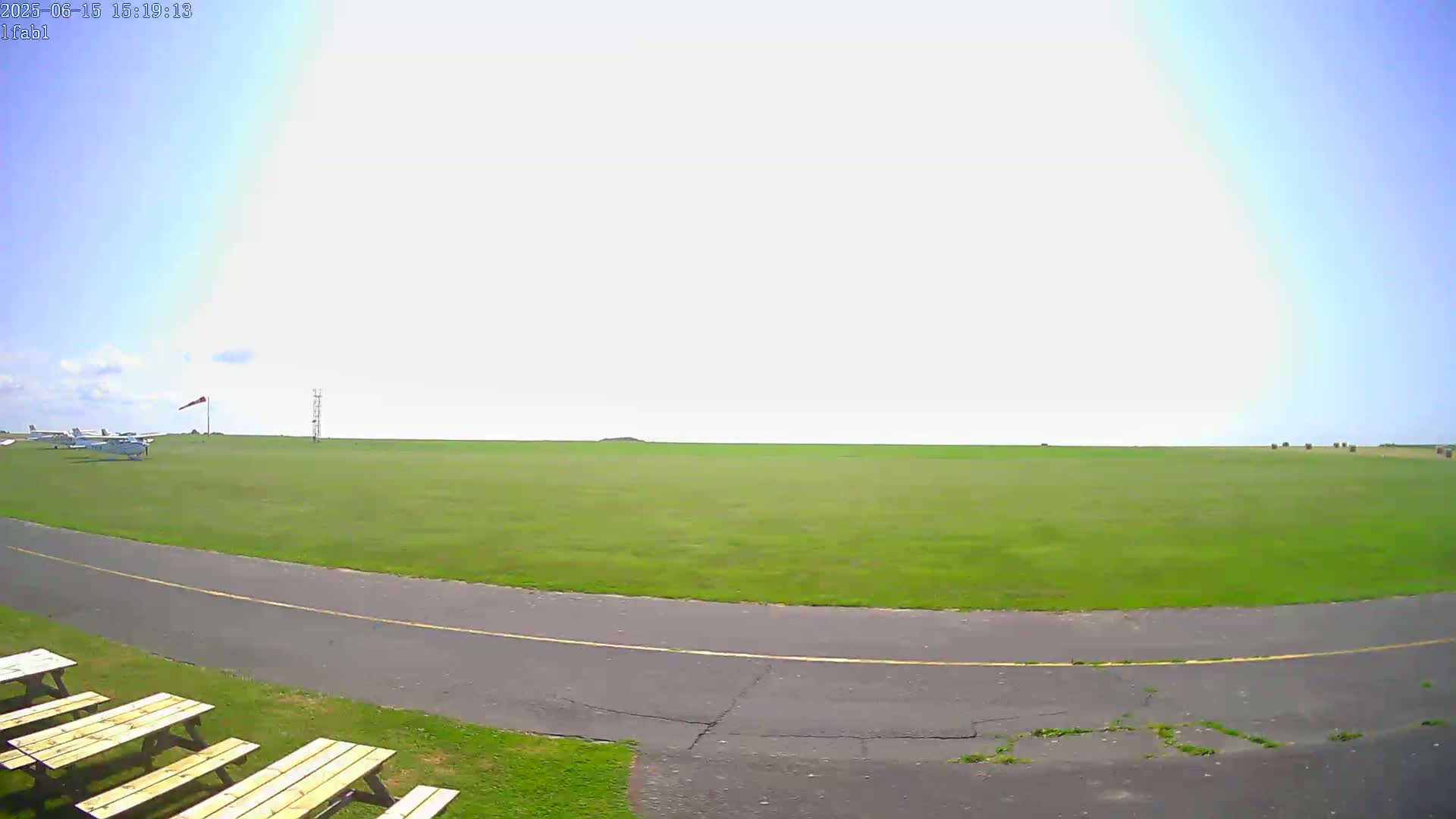 A grassy airfield with a paved taxiway, a few small planes, and picnic tables is visible under a mostly sunny sky.