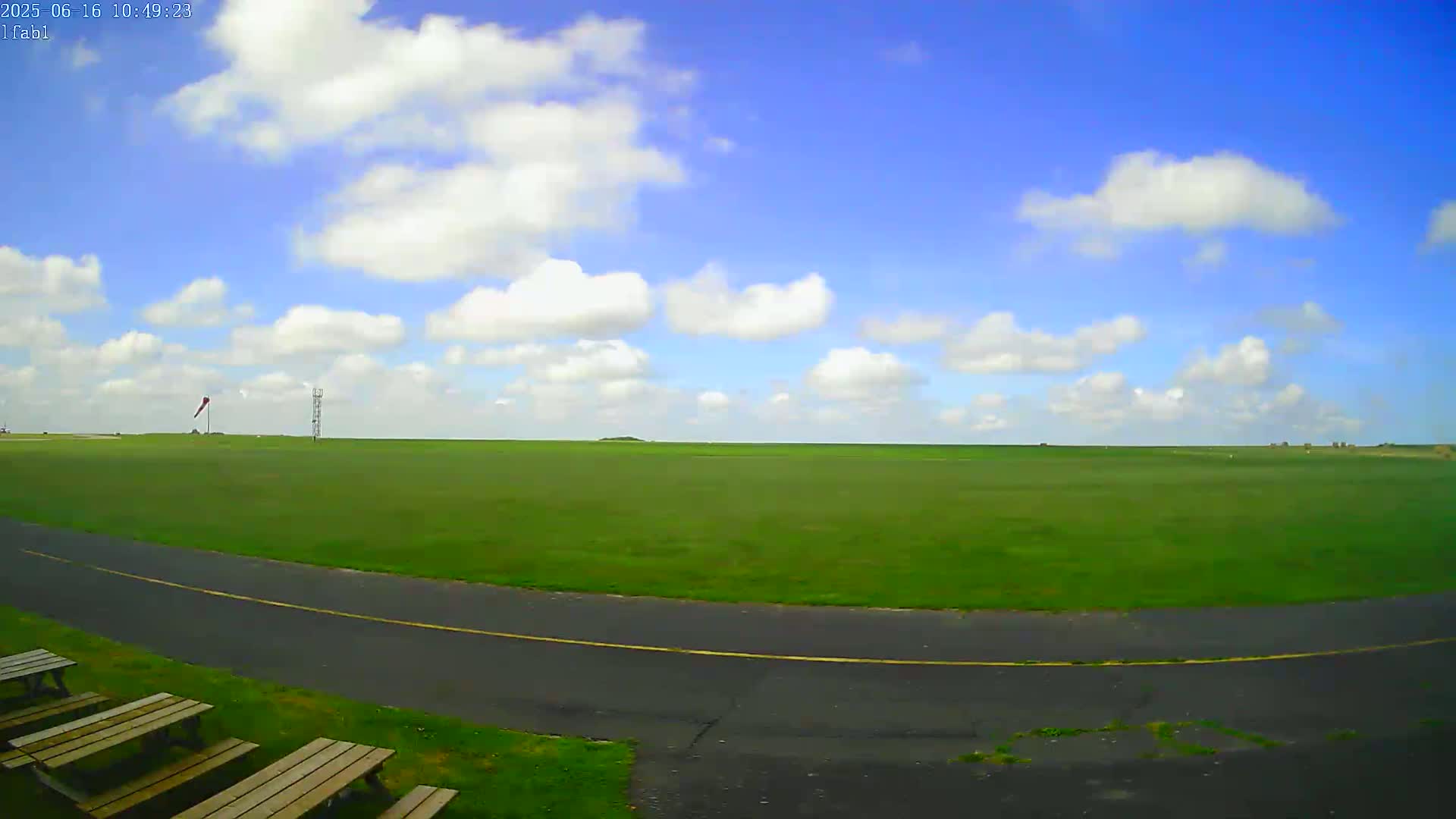 A wide, grassy field with a paved roadway curving through it, under a mostly sunny sky with scattered cumulus clouds.
