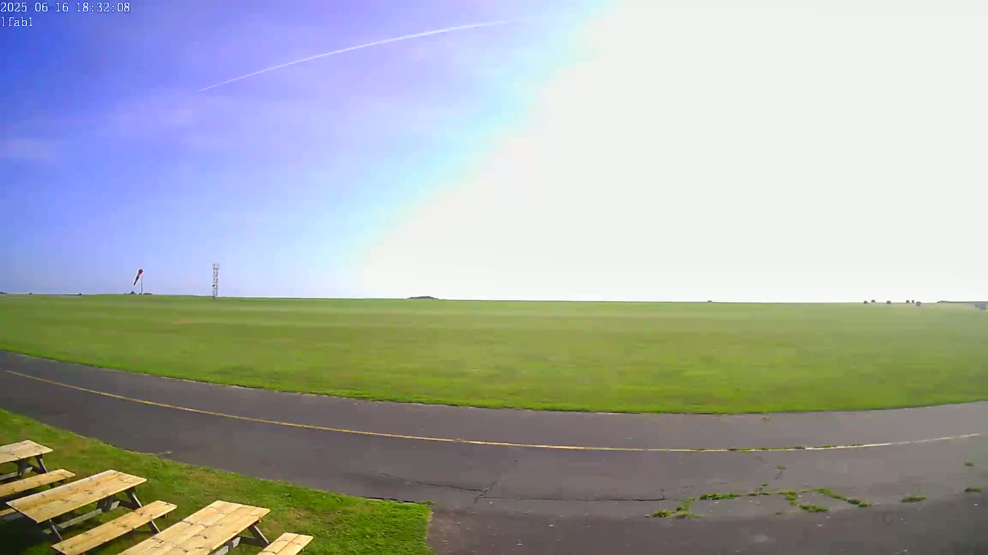 A grassy airfield under a mostly clear, bright sky, with a paved roadway curving through the foreground and picnic tables to the left.