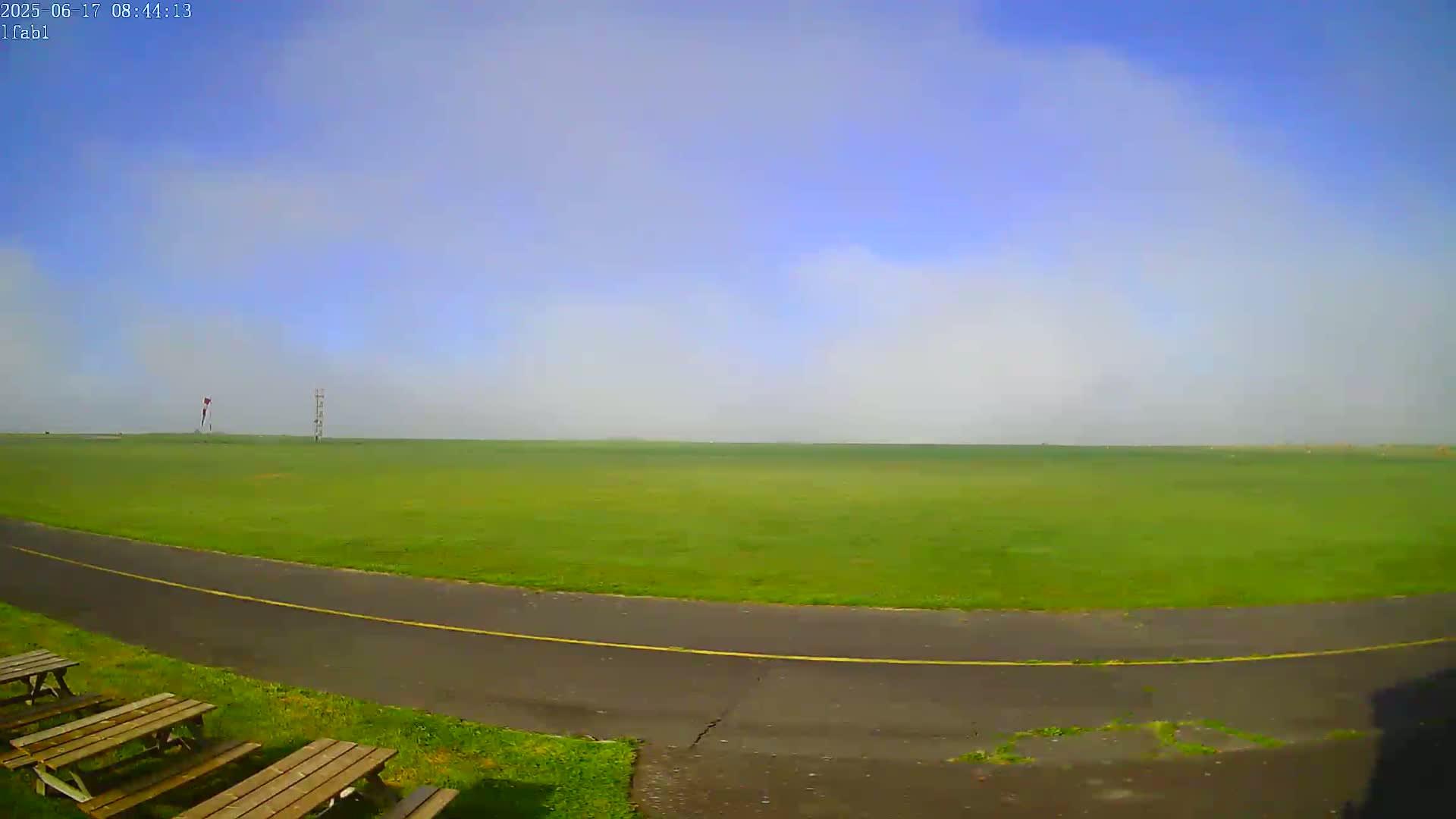 A wide, grassy field under a mostly clear, slightly hazy sky is viewed from a low vantage point, with a paved path curving in the foreground and picnic tables to the left.