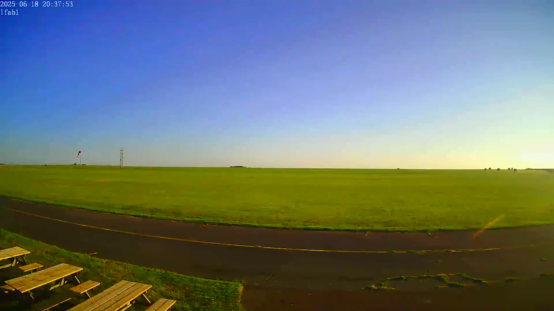 A wide, flat expanse of bright green grass under a clear blue sky is bordered by a dark asphalt path, with several wooden picnic tables in the foreground.