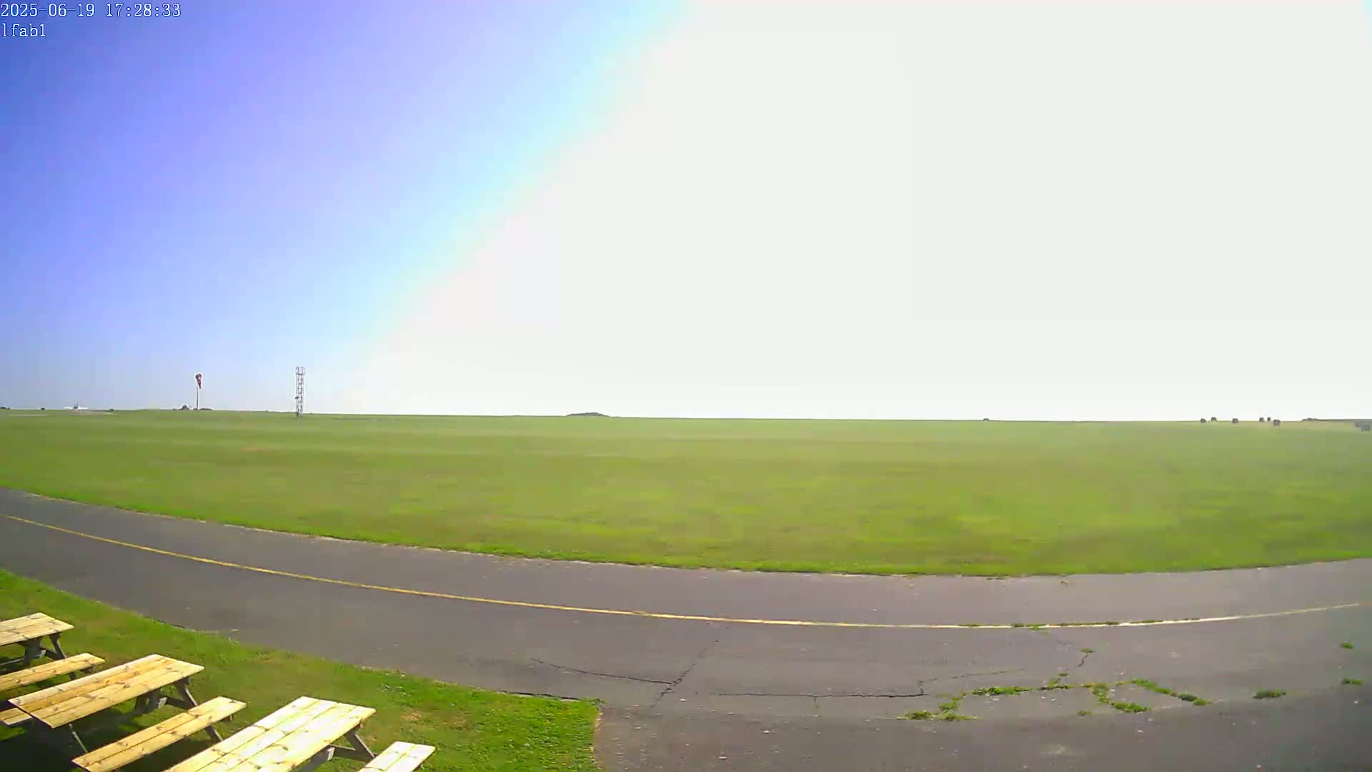 A vast, grassy airfield under a clear, bright sky is visible from a low vantage point, with a paved roadway curving into the foreground, where several picnic tables sit.