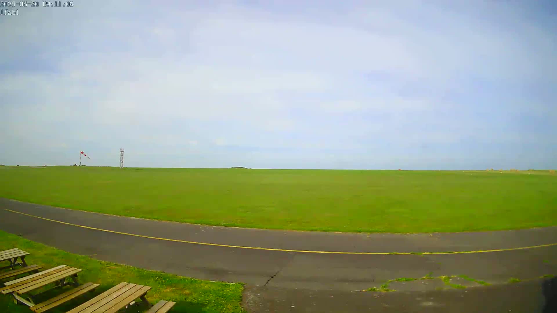 A mostly sunny day reveals a large, grassy field with a paved roadway curving through it, a windsock, and several picnic tables in the foreground.