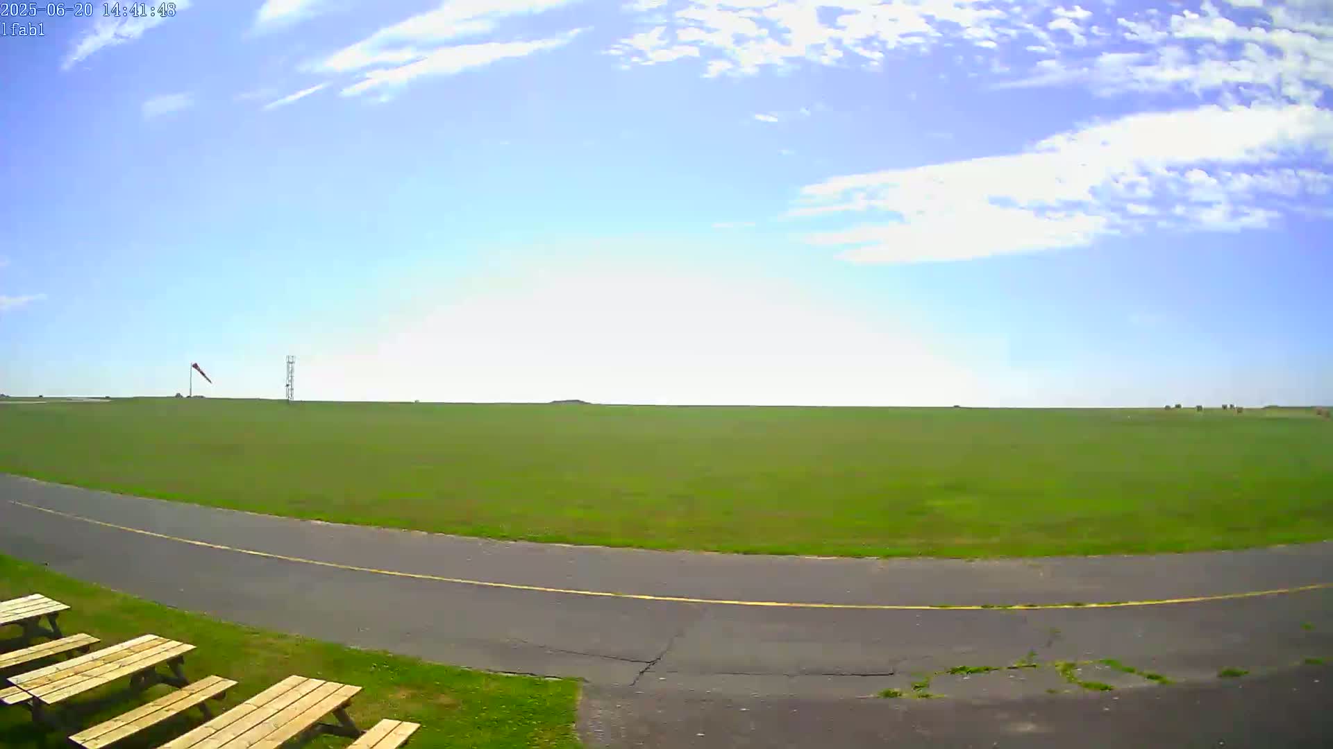 A mostly sunny day shows a grassy airfield with a paved taxiway curving toward the viewer, picnic tables nearby, and a windsock in the distance.