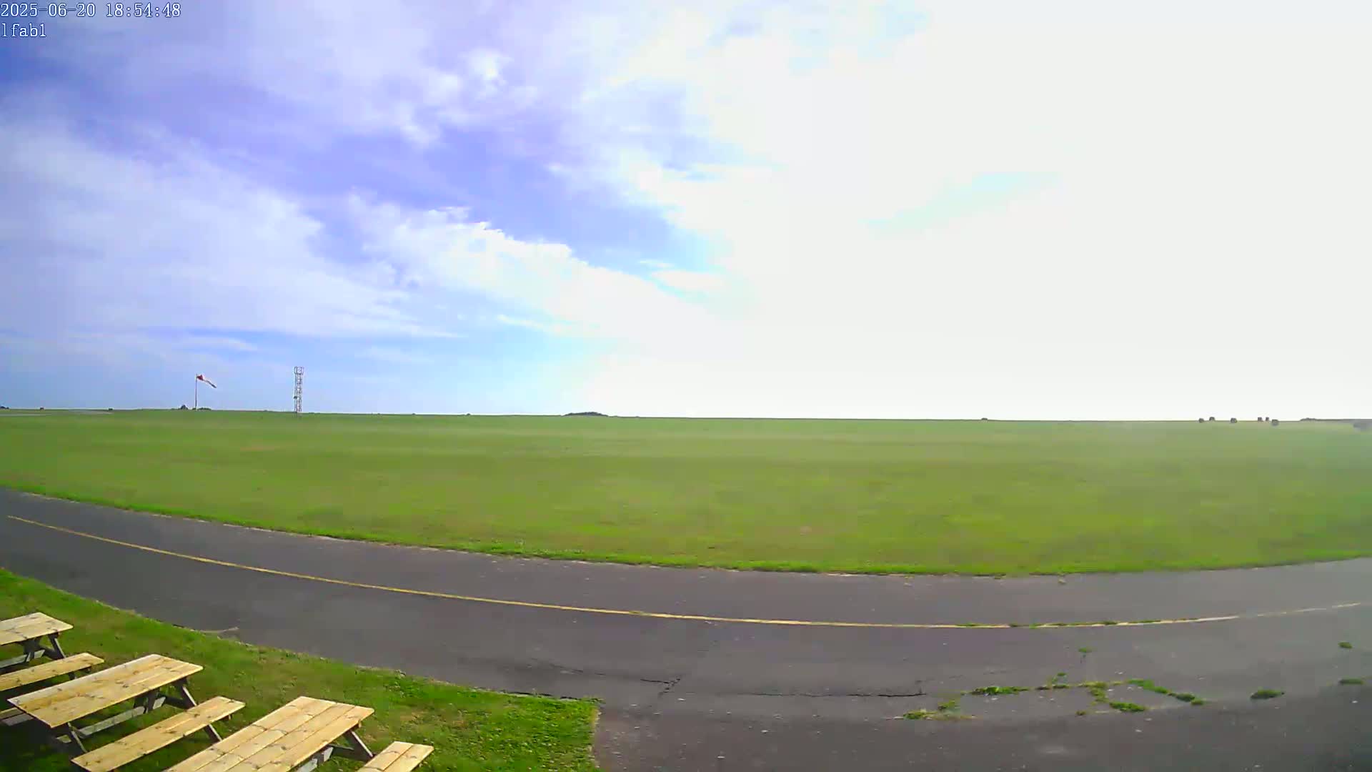 A wide, grassy field under a partly cloudy sky is seen from a low vantage point, with a paved path curving in the foreground and several wooden picnic tables to the left.