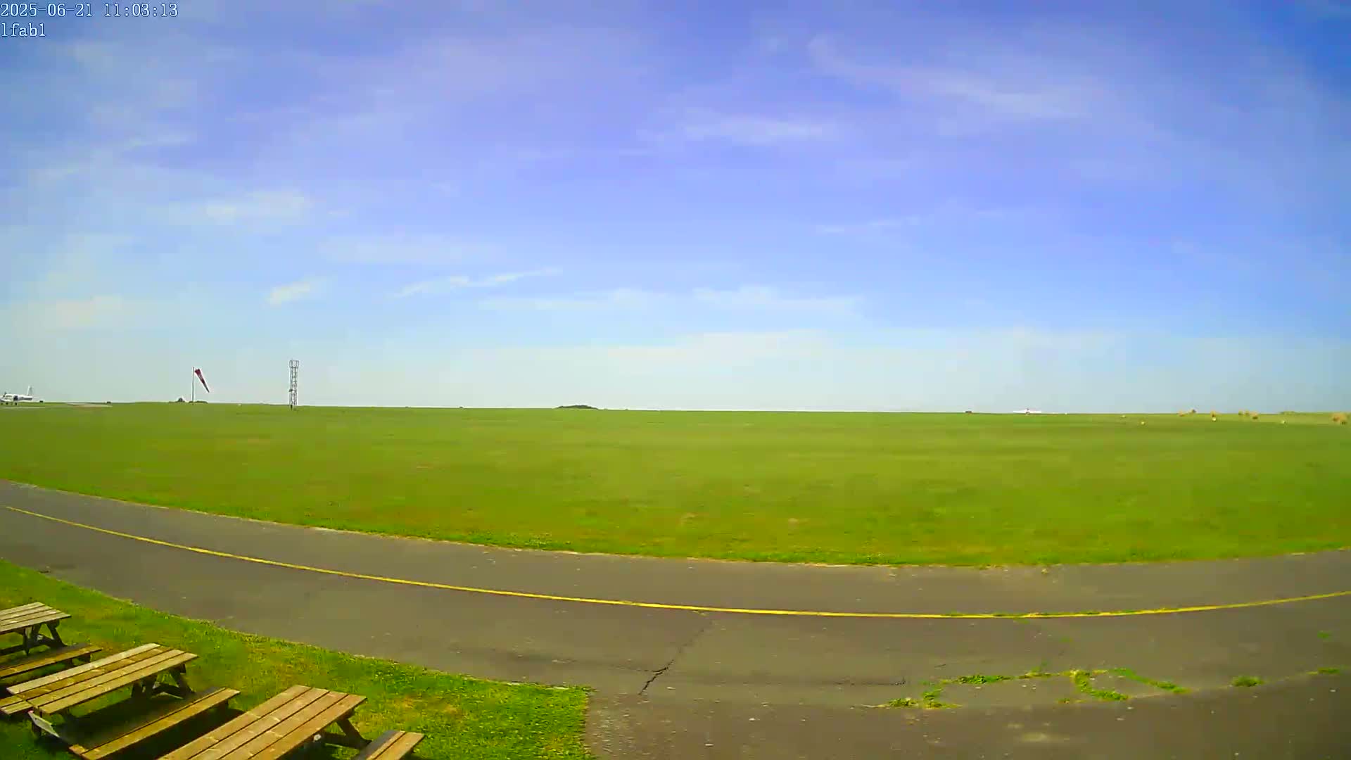 A mostly sunny day reveals a large expanse of green grass, a paved path, and several picnic tables near a small airfield.