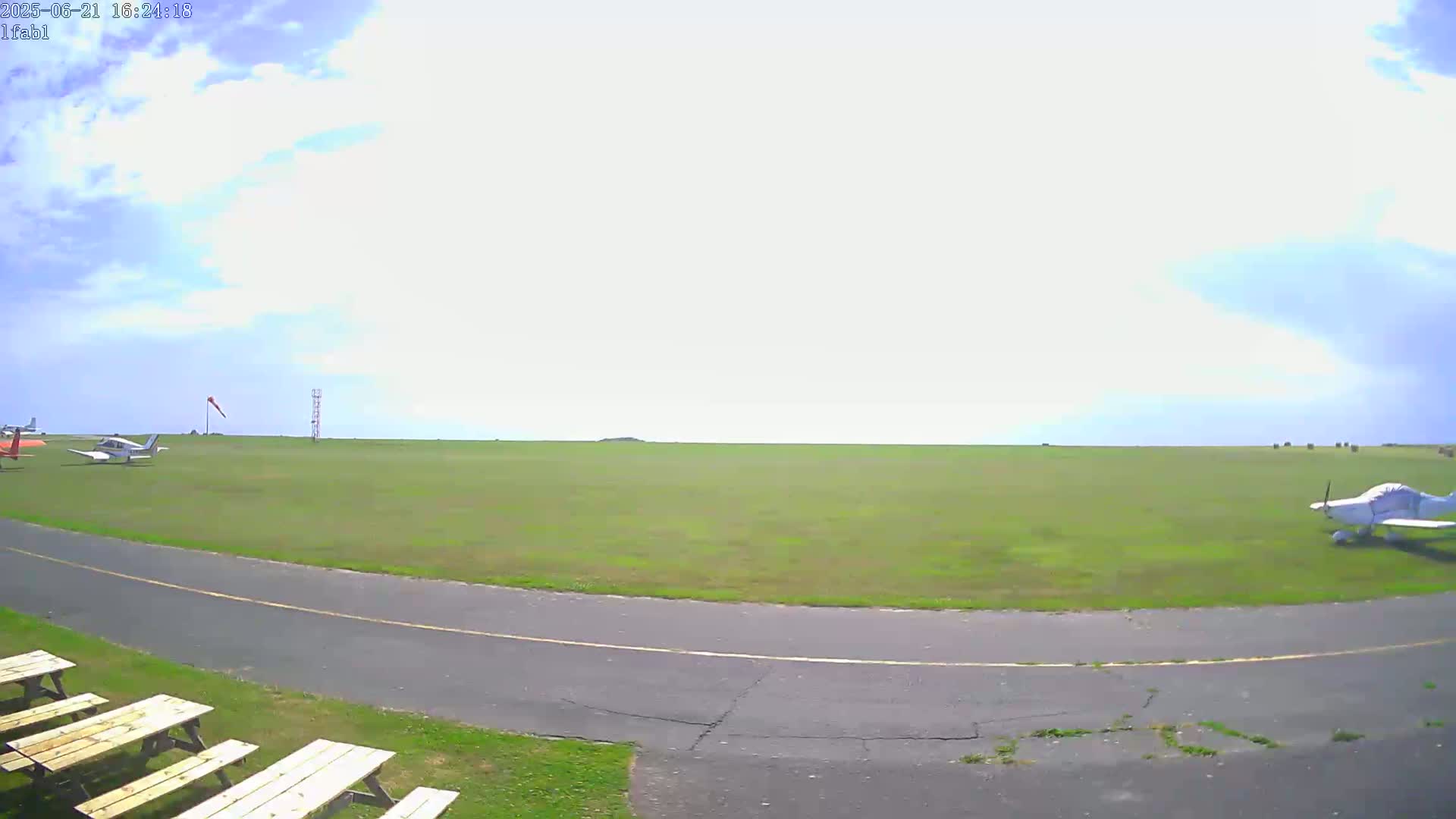 A grassy airfield under a partly cloudy sky shows several small airplanes parked on the tarmac near a paved roadway and picnic tables.