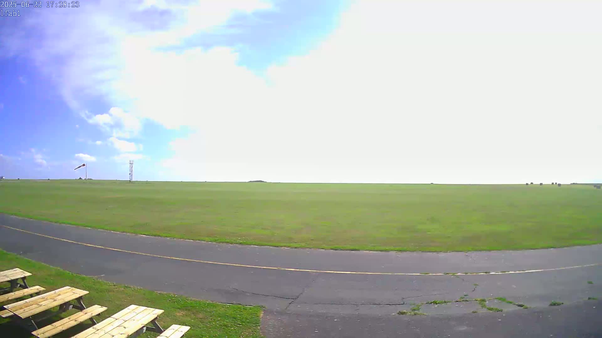 A mostly sunny day shows a grassy airfield with a paved taxiway, a windsock, and picnic tables.