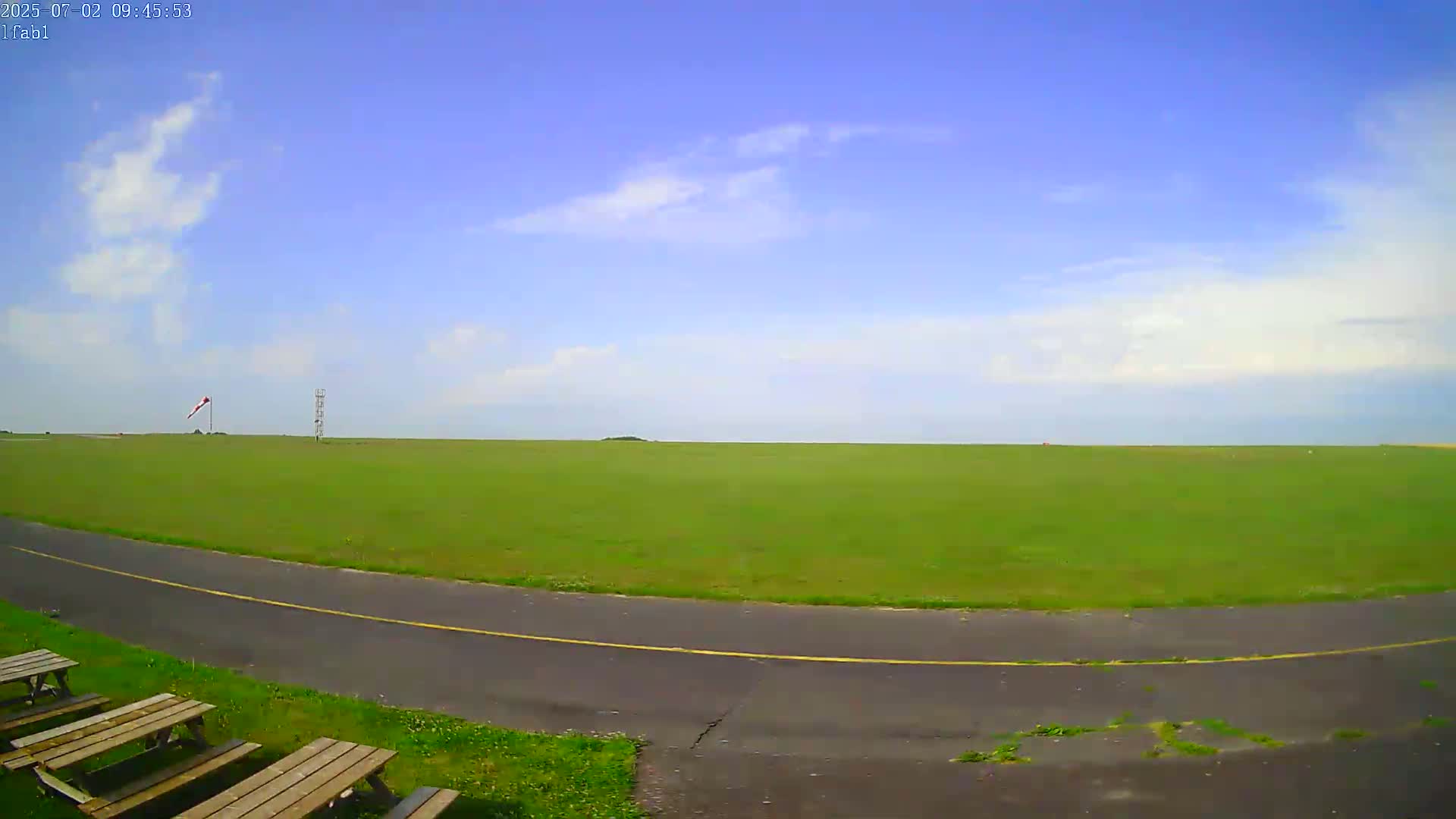 A grassy airfield under a mostly clear blue sky with a few scattered clouds is visible, alongside a paved roadway and several picnic tables.