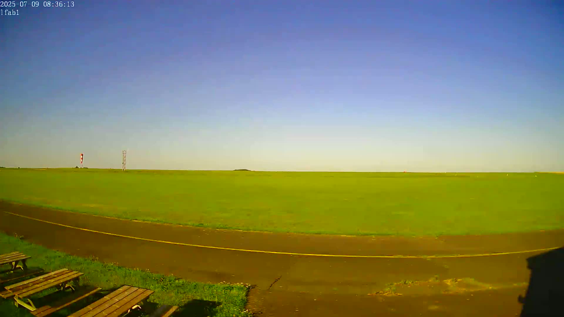 A wide, grassy field under a clear blue sky is visible, with a paved path and picnic tables in the foreground.