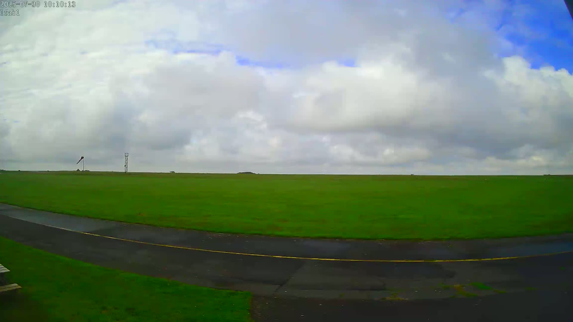 A mostly cloudy sky overlooks a large expanse of bright green grass with a paved road curving through the bottom.