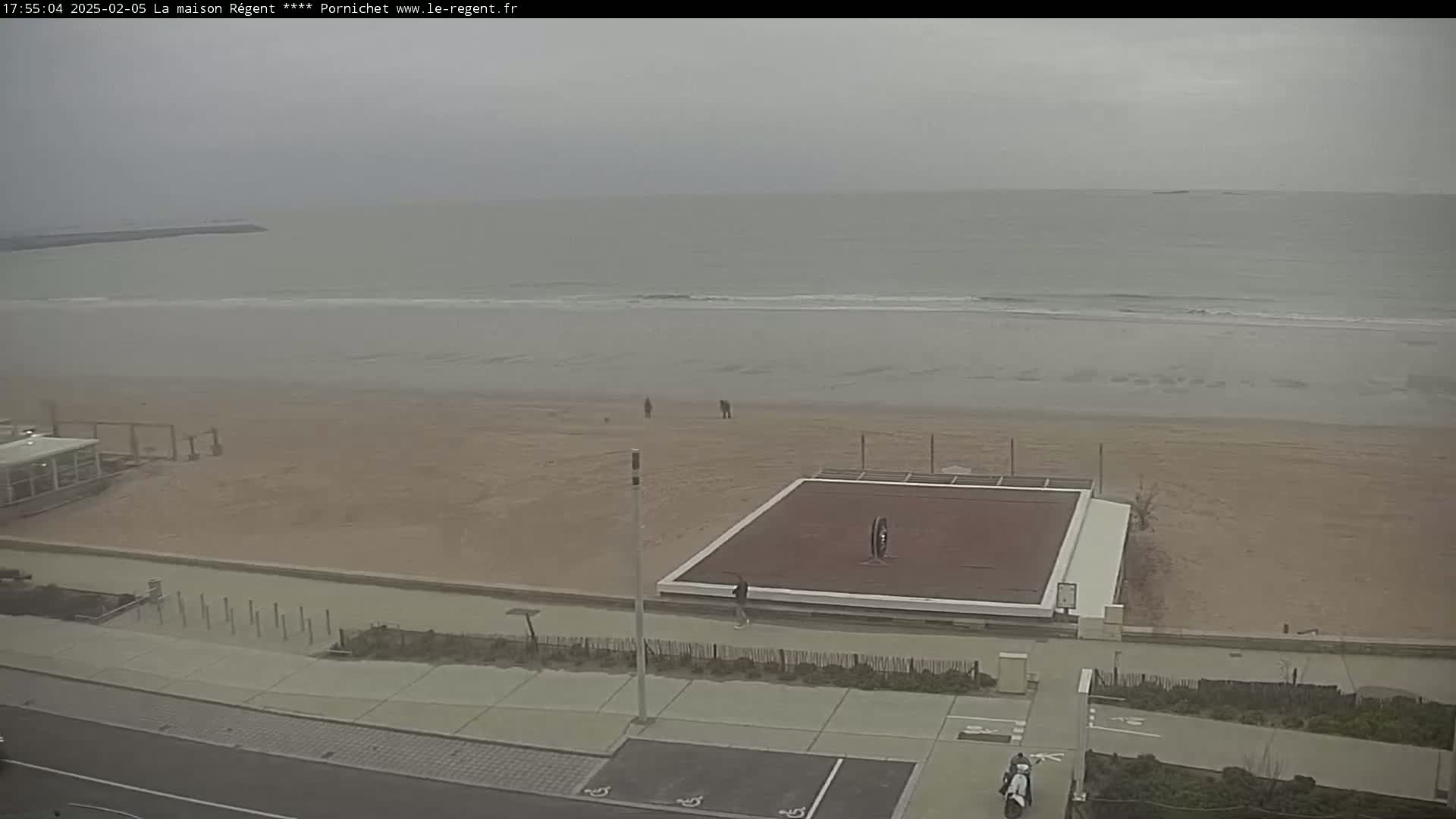 A mostly empty sandy beach stretches to a calm, gray sea under an overcast sky, with a few people strolling along the shore and a volleyball court near the boardwalk.