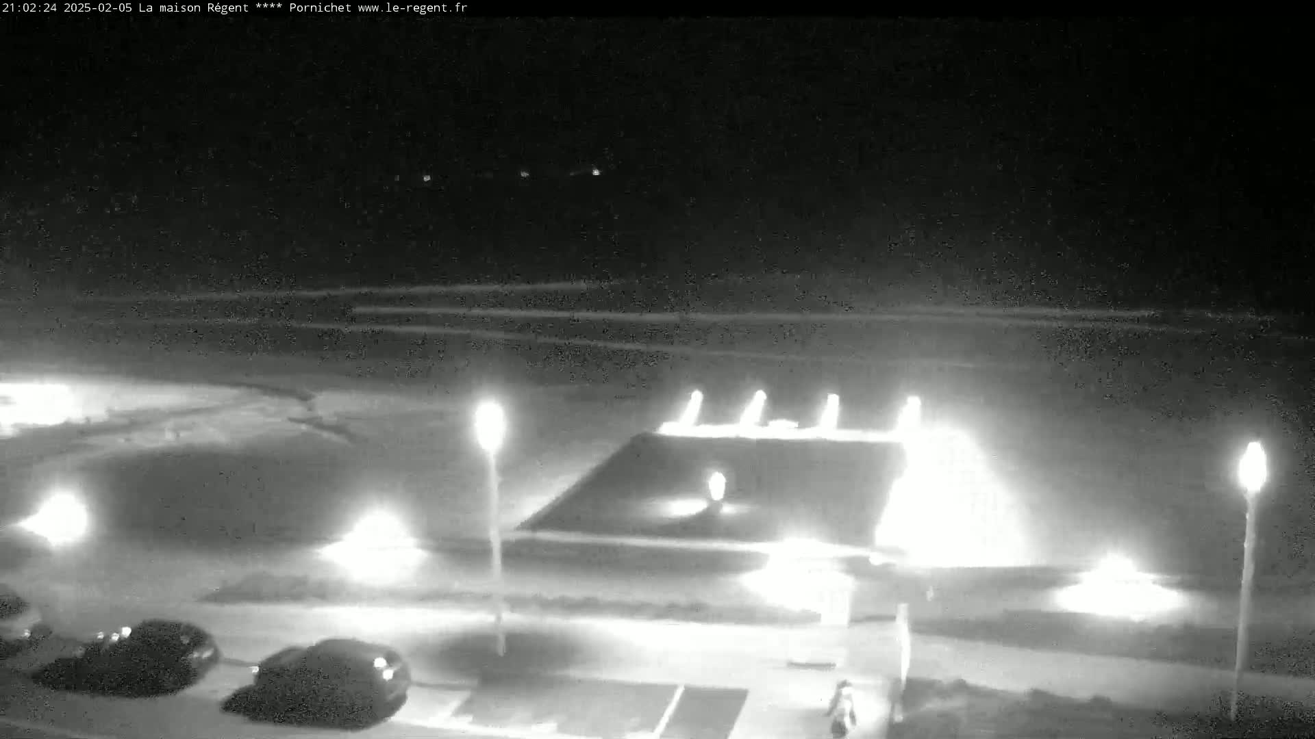 A nighttime, low-light view of a beach area with several cars parked, lamps illuminating a paved area, and a person walking near some buildings.