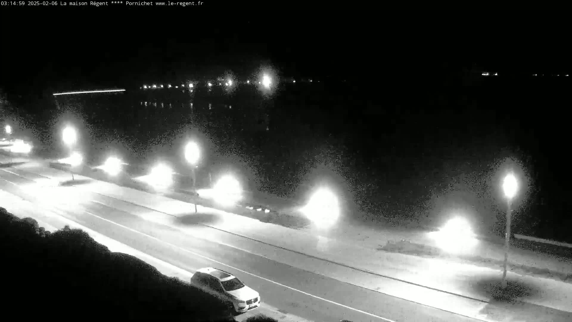 A nighttime, high-angle view of a road next to a body of water, illuminated by streetlights, with a single car visible on the road.