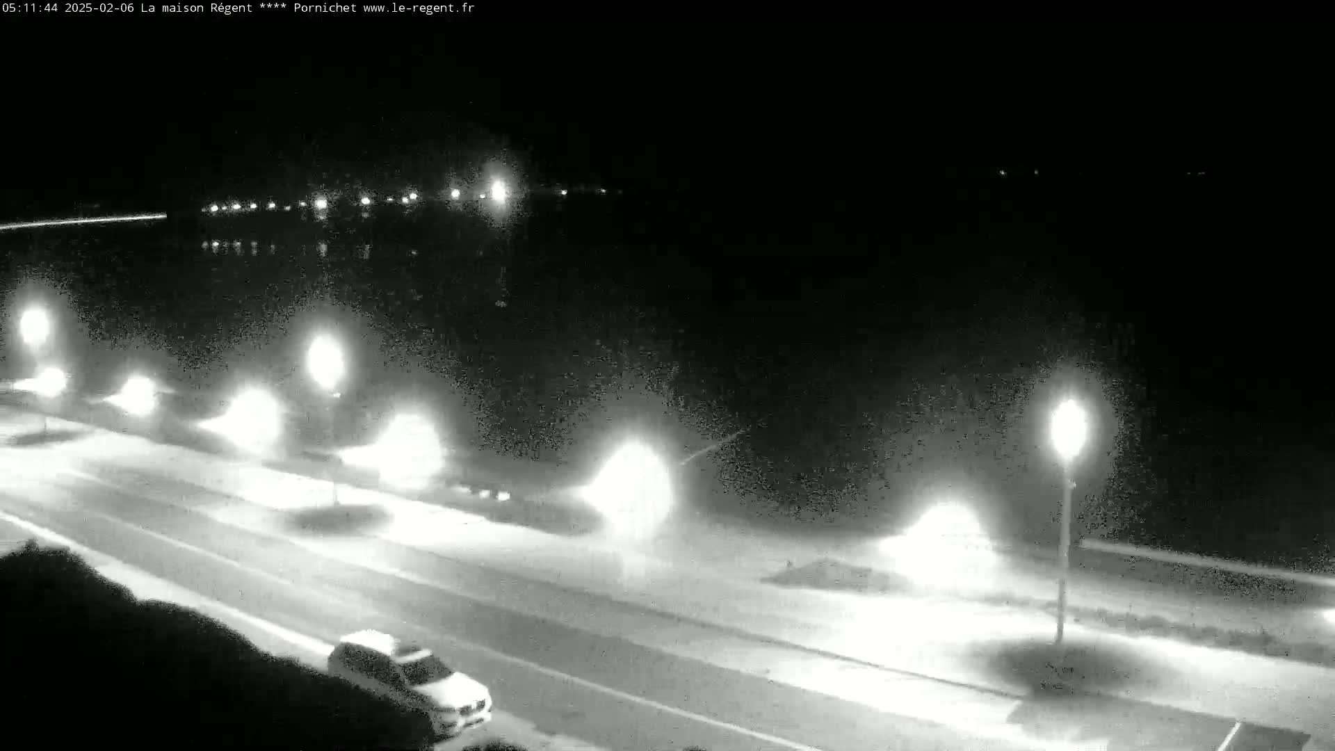 A nighttime, monochrome view shows a road with cars and streetlights next to a dark body of water.