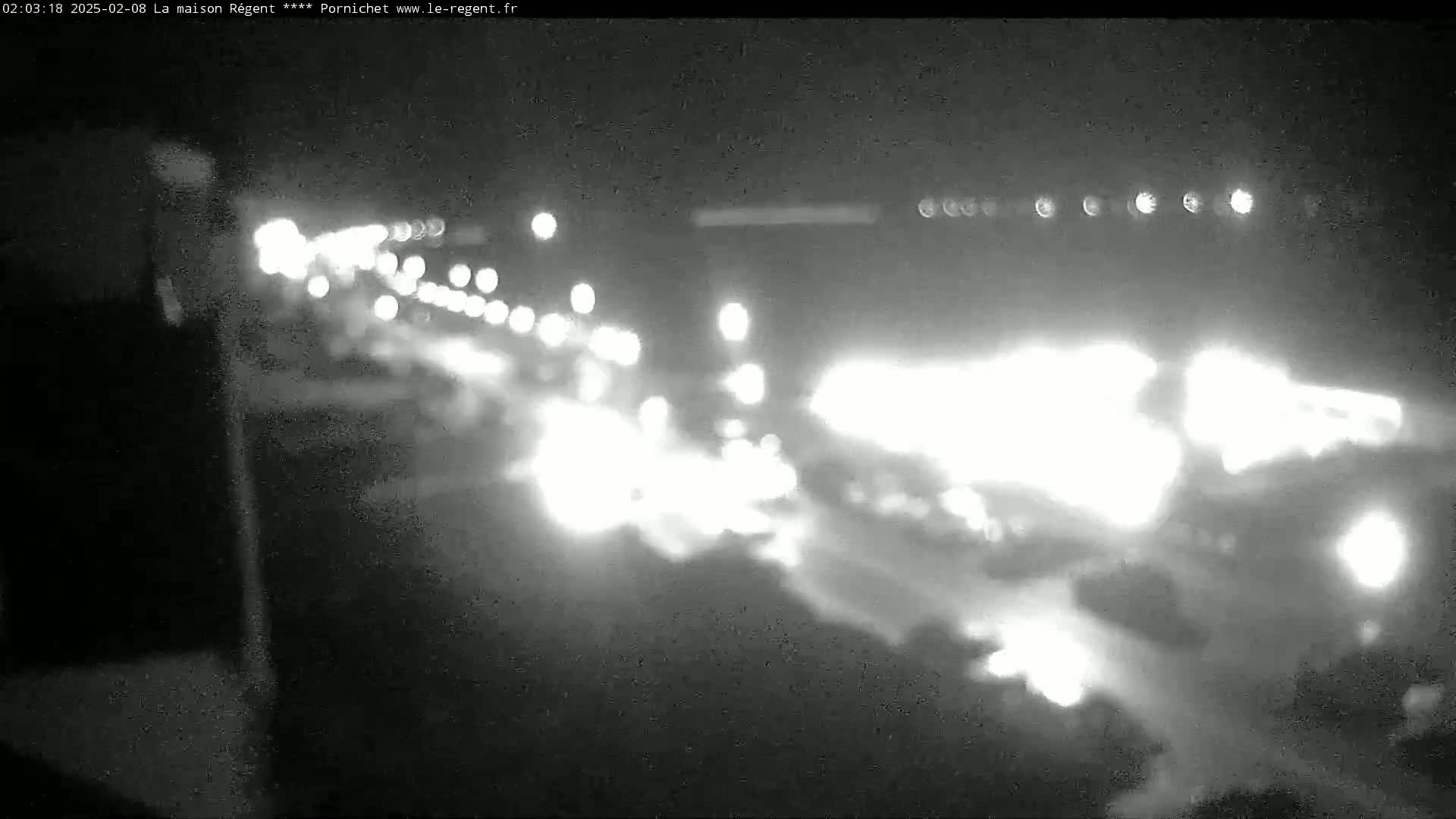 A nighttime, blurry, black and white view of a road with bright lights from vehicles and buildings in the distance.