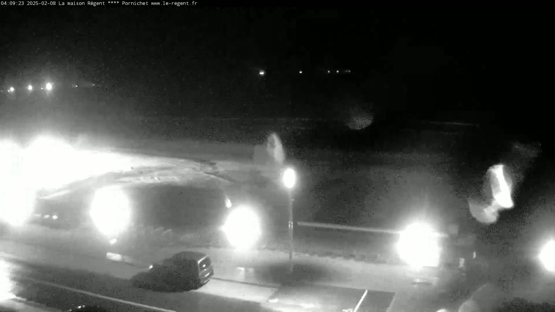 A nighttime, low-light, grainy security camera image shows several vehicles driving on a road with a single lamppost visible, under what appears to be clear skies.