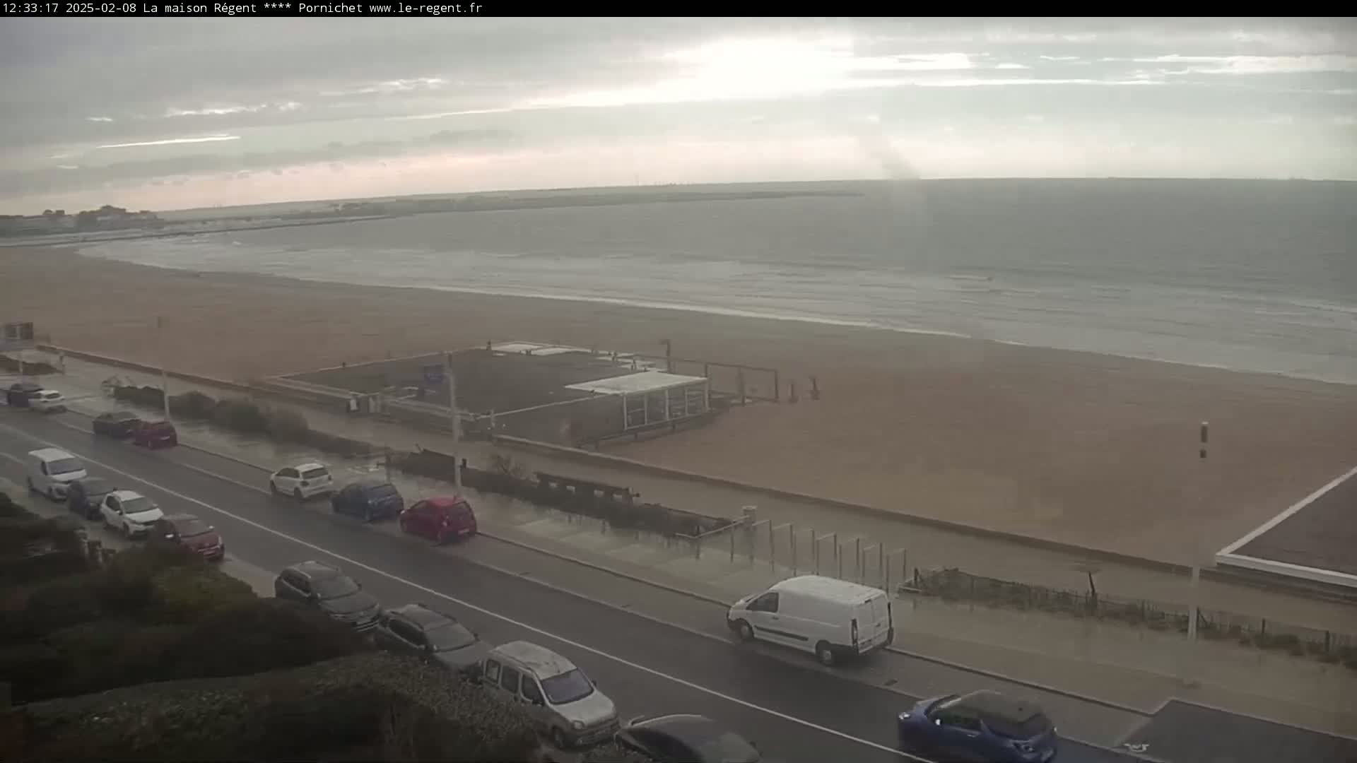 A mostly cloudy day shows a wet beach with small waves and several cars parked along a seaside road.