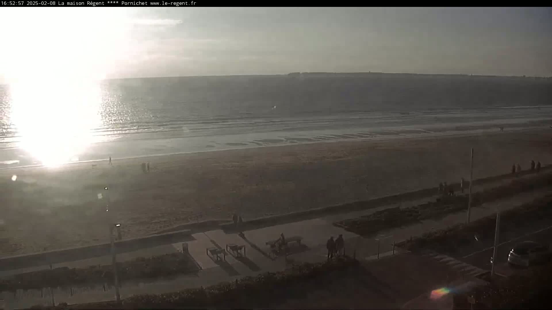A mostly sunny day shows a wide sandy beach with calm waves and a few people sparsely scattered along the shore and boardwalk.