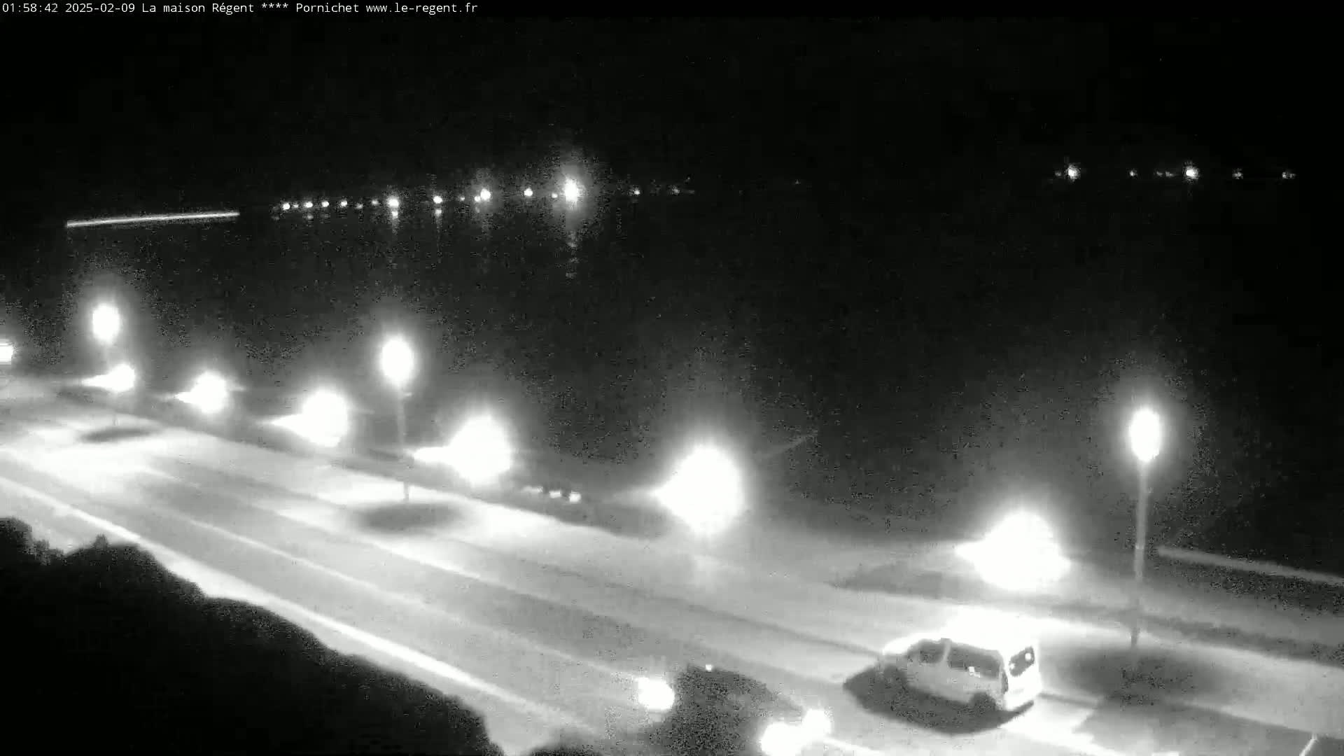 A nighttime, overhead view shows a road next to a body of water, with cars driving on the road and lights illuminating the scene.