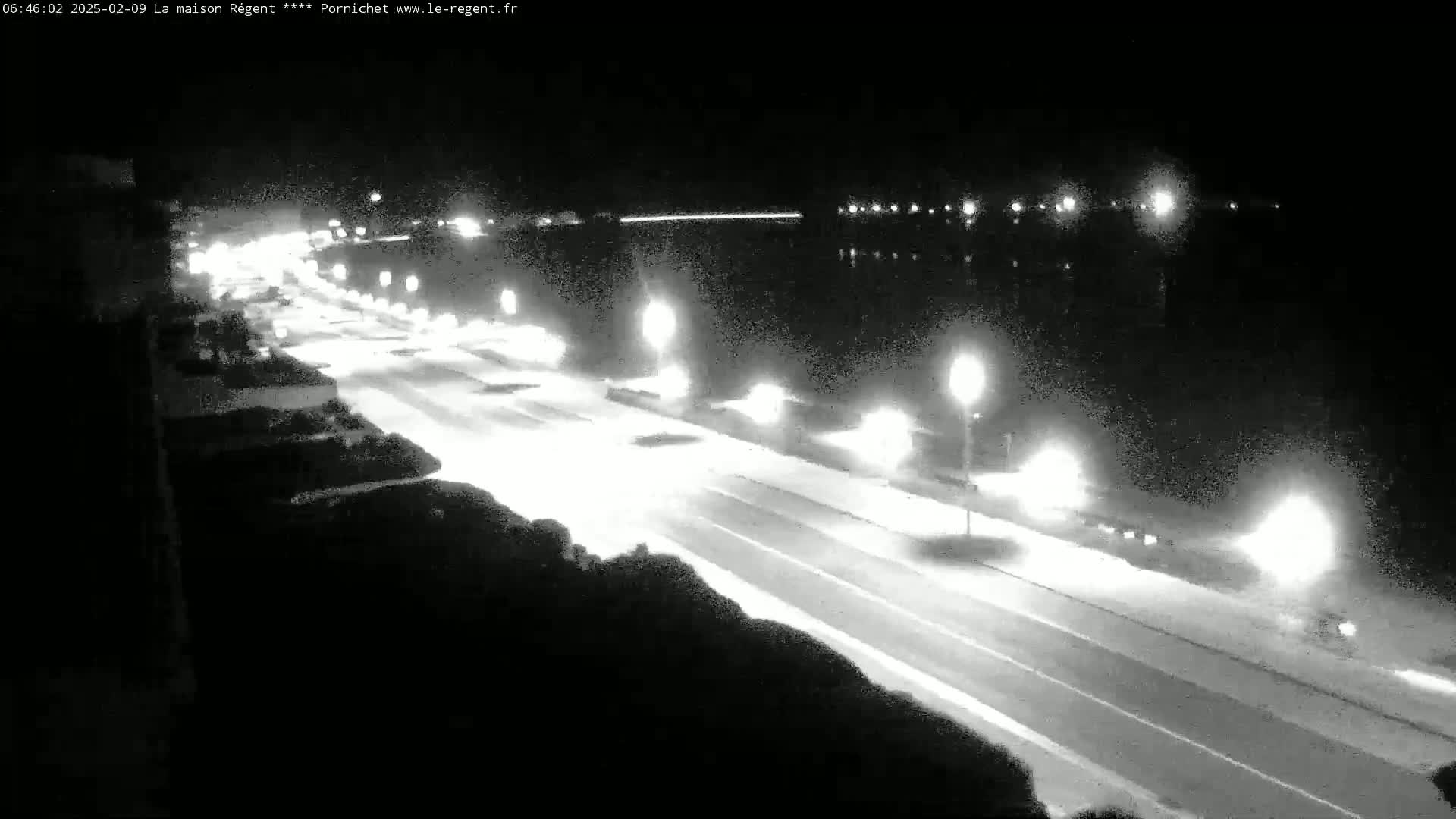 A nighttime, high-angle view shows a brightly lit roadway next to a body of dark water, with vehicles traveling on the road.