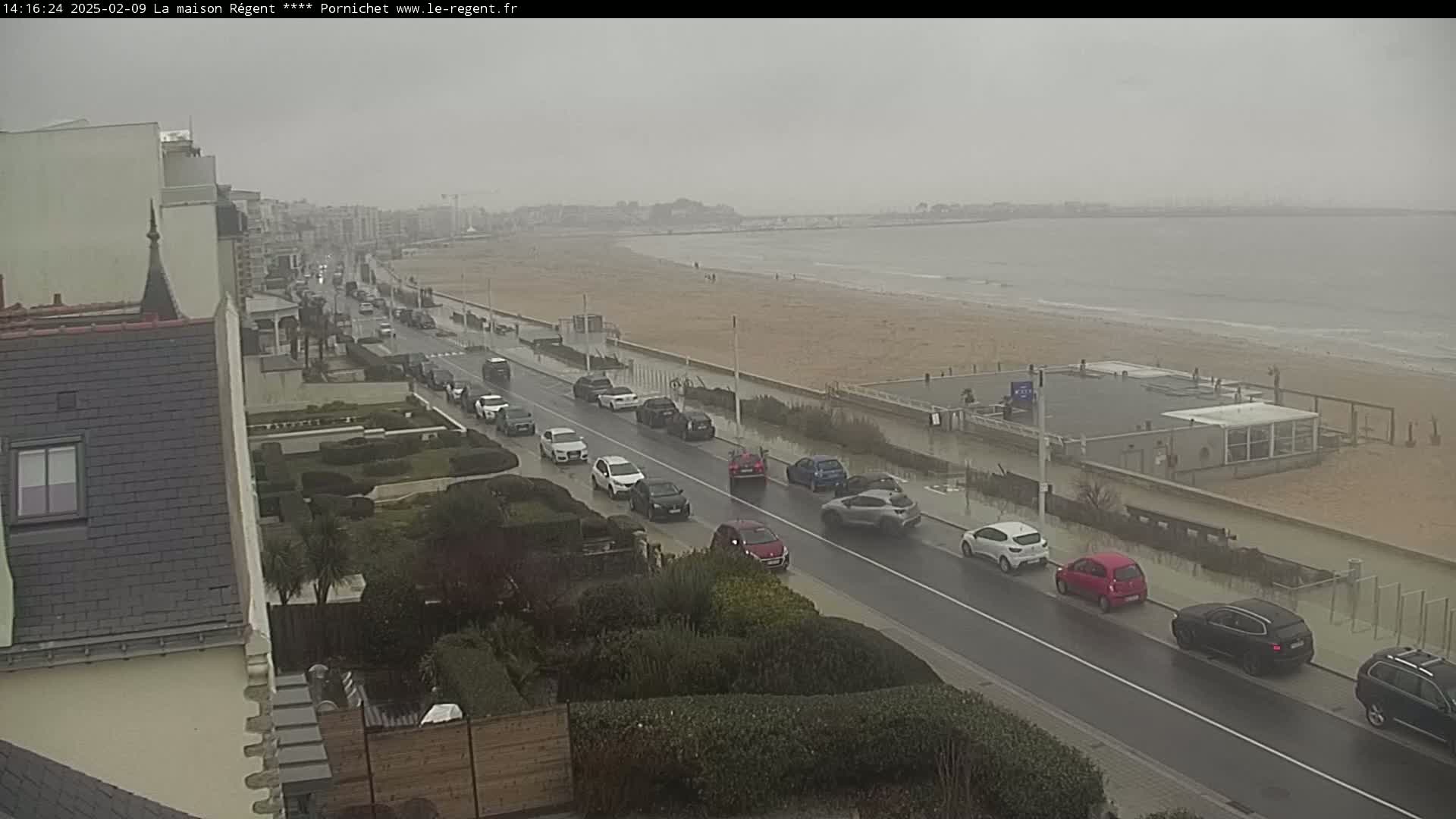 A wet, overcast day shows a seaside road with cars driving past a beach and buildings.