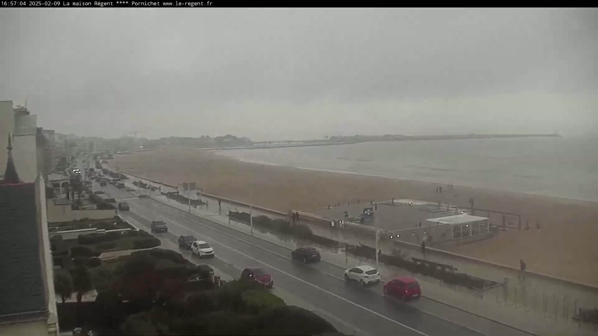 A rain-swept coastal road with cars driving past a wide sandy beach and calm ocean under an overcast sky.
