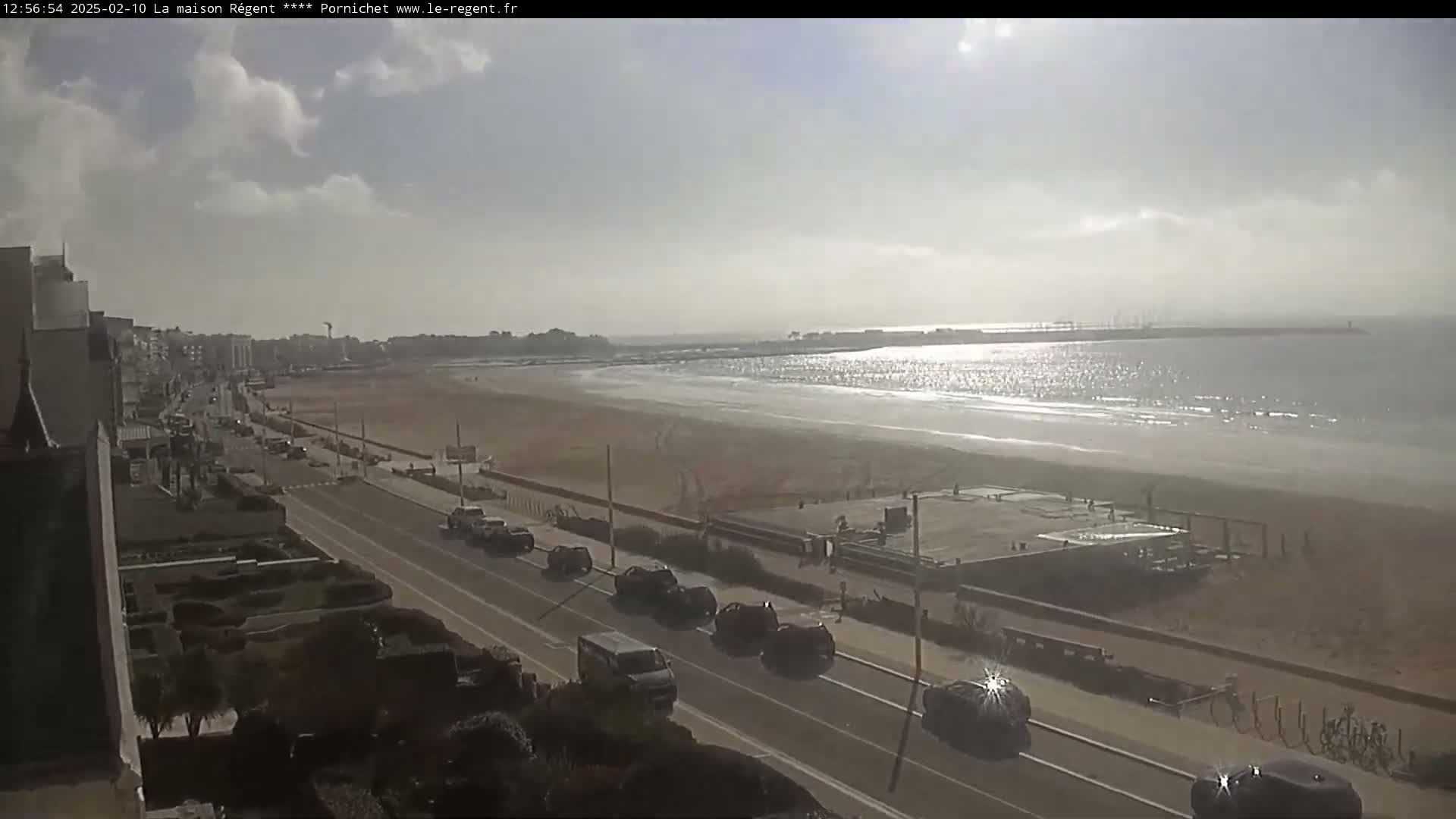 A mostly sunny day shows a long sandy beach with calm waves, a road with several cars driving along the beachfront, and buildings in the distance.
