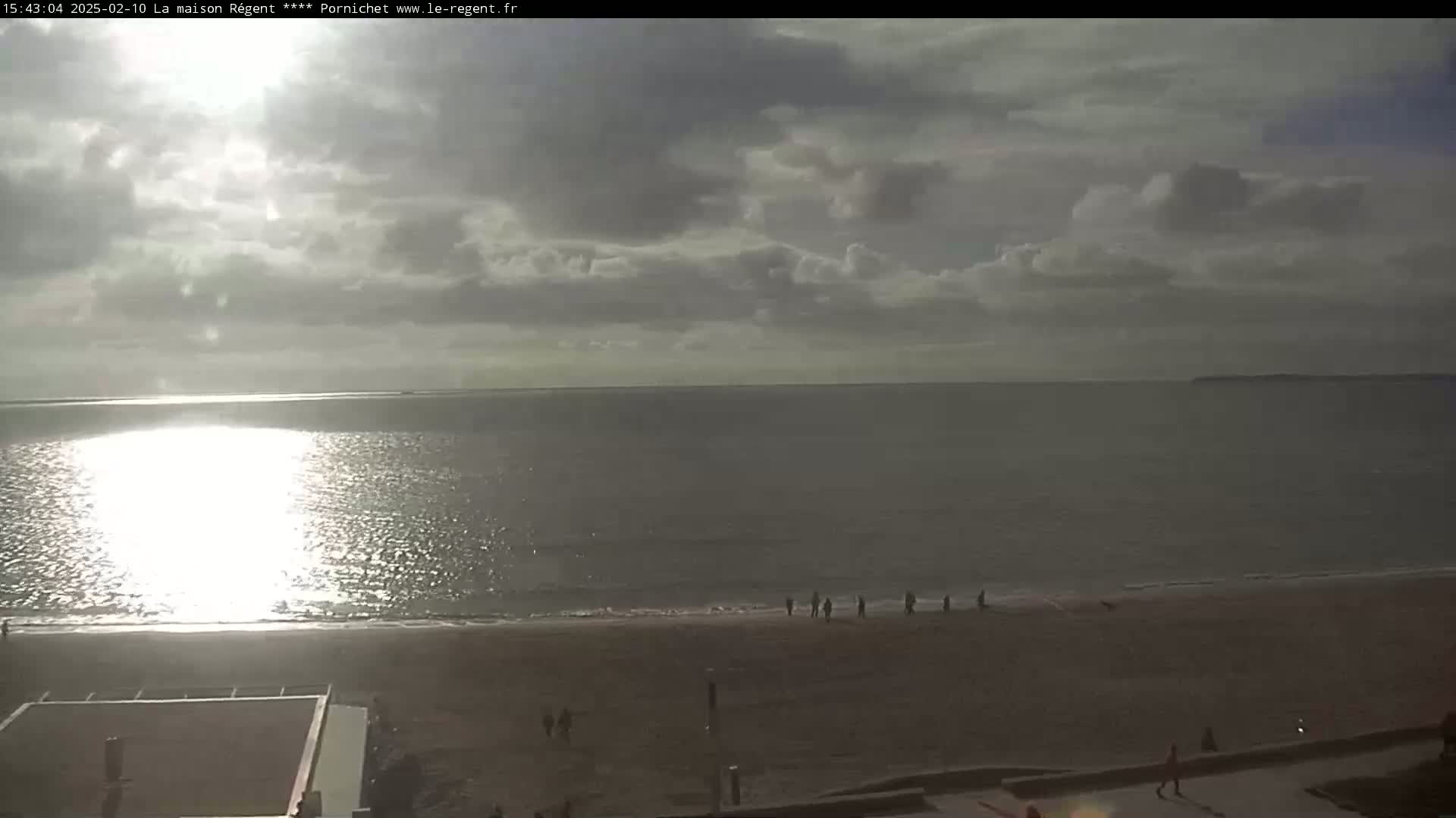 A mostly cloudy day shows a calm ocean reflecting sunlight, with a long sandy beach and a few people walking along the shore.