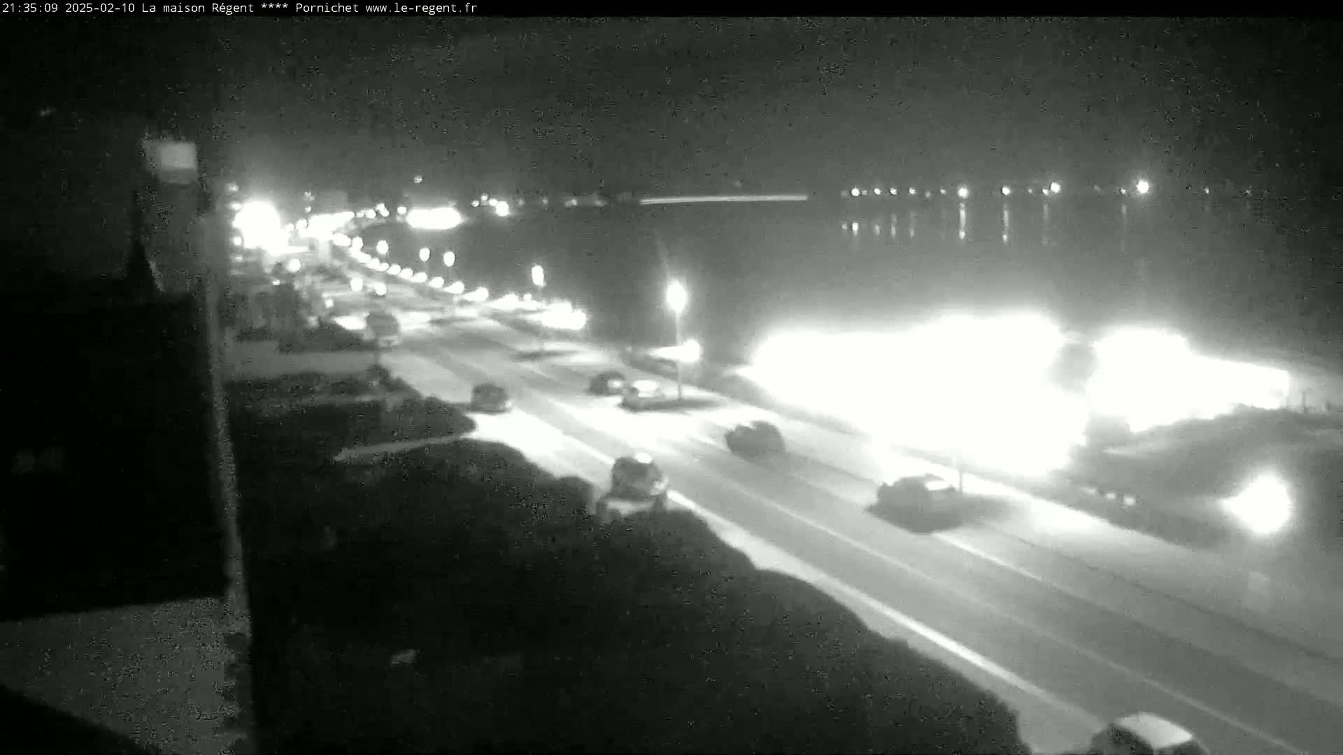A nighttime, high-angle view shows a road with cars driving alongside a body of water under a dark sky.