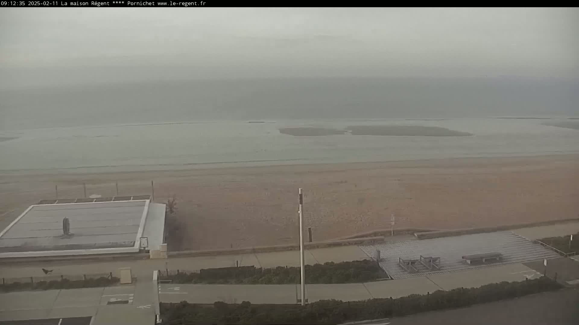 A hazy, overcast day reveals a wide sandy beach, calm ocean water, and a paved walkway leading to a beachfront structure.