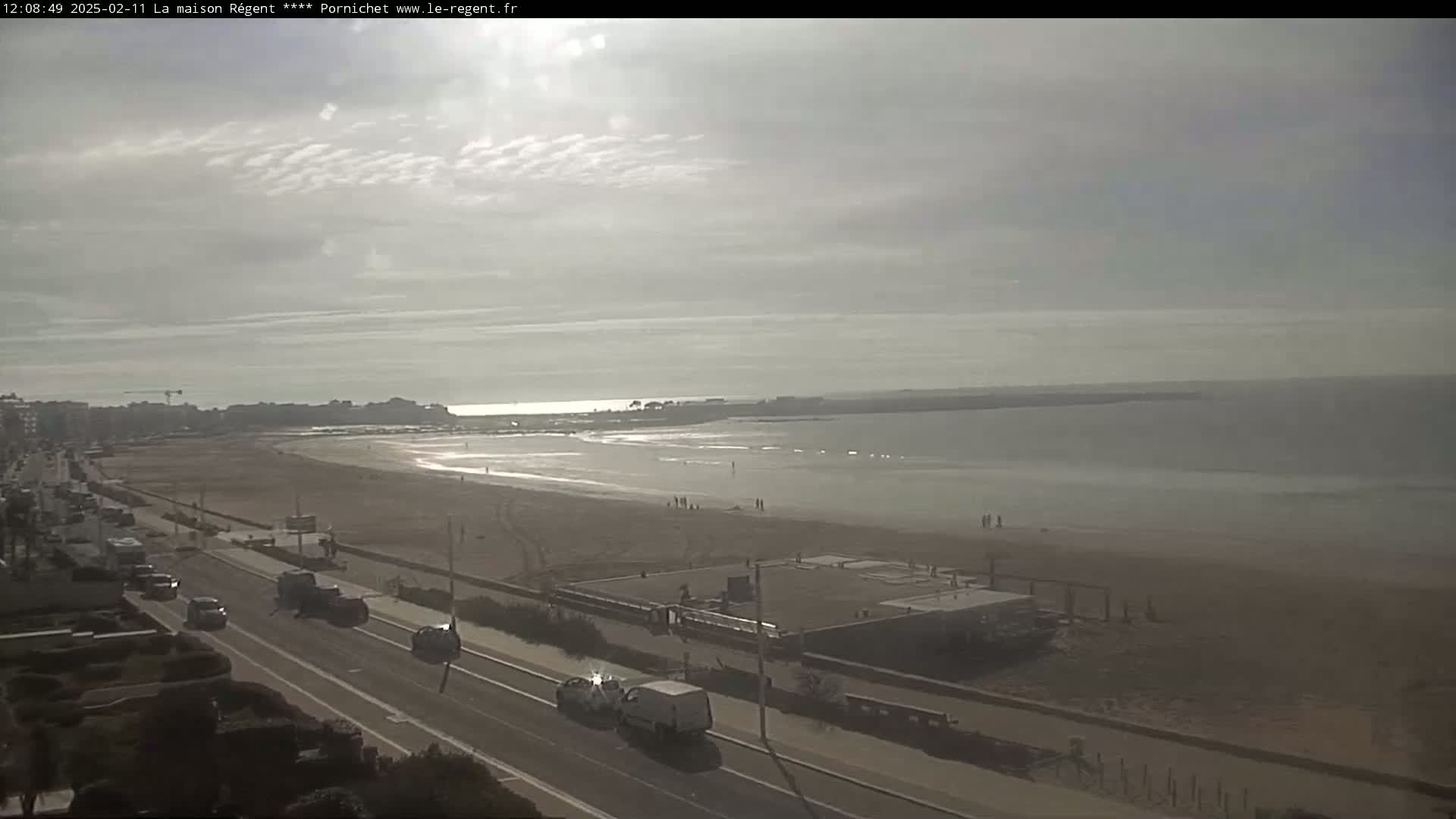 A mostly sunny day reveals a beach with a few people, a road with cars, and buildings in the distance.