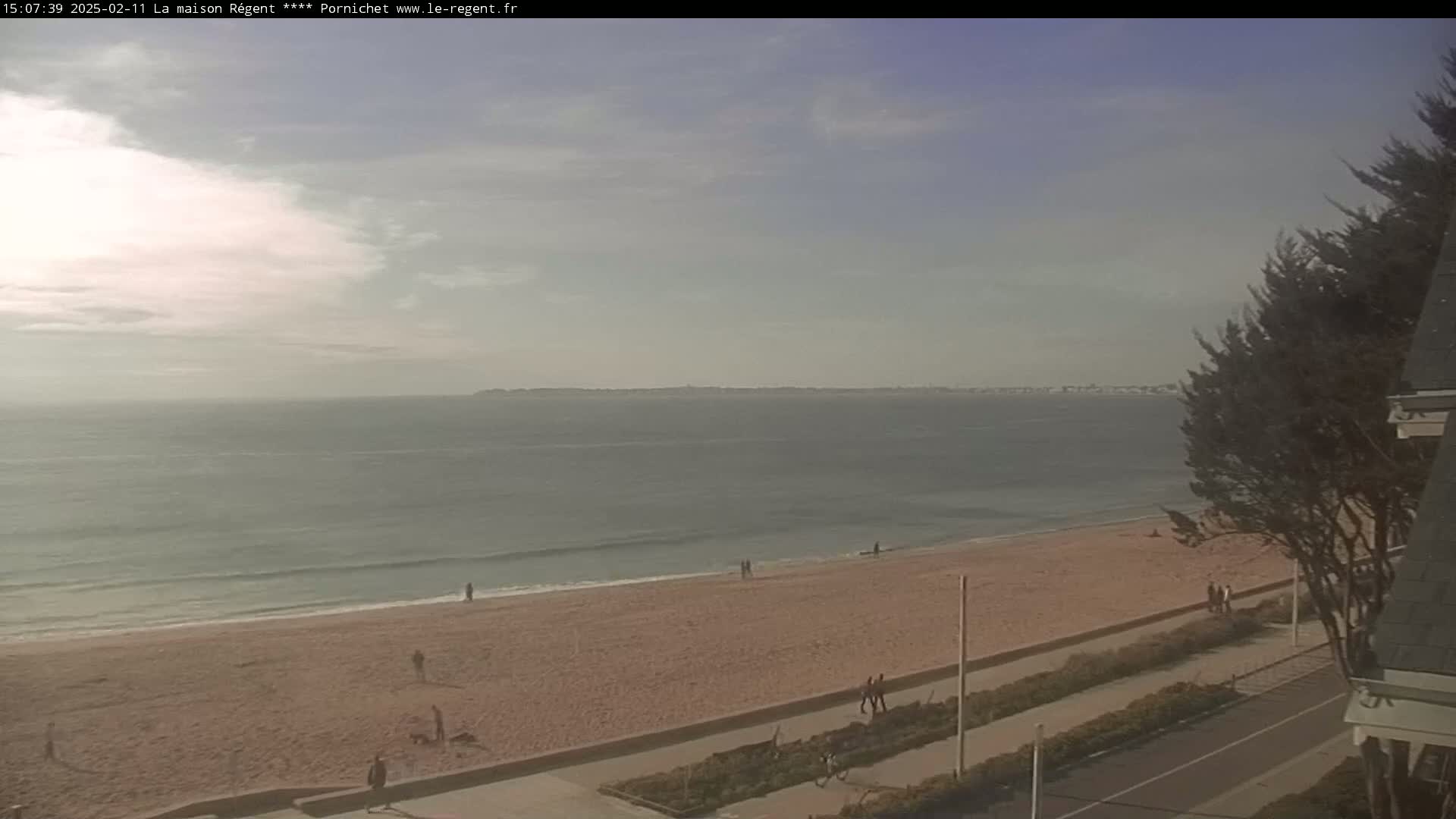 A mostly cloudy day shows a sandy beach with a few people, and calm ocean waves lapping the shore, with a distant landmass visible across the water.