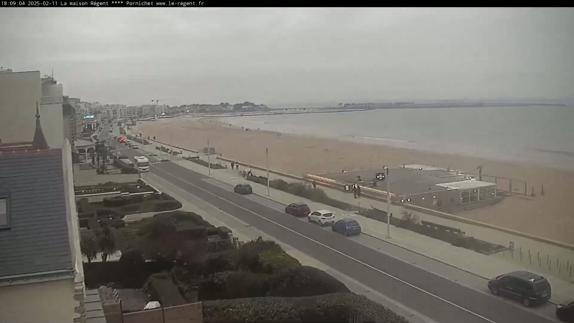 A seaside town with a wide sandy beach, calm water, and overcast skies is seen from an elevated perspective, showing cars parked along a road bordering the beach.