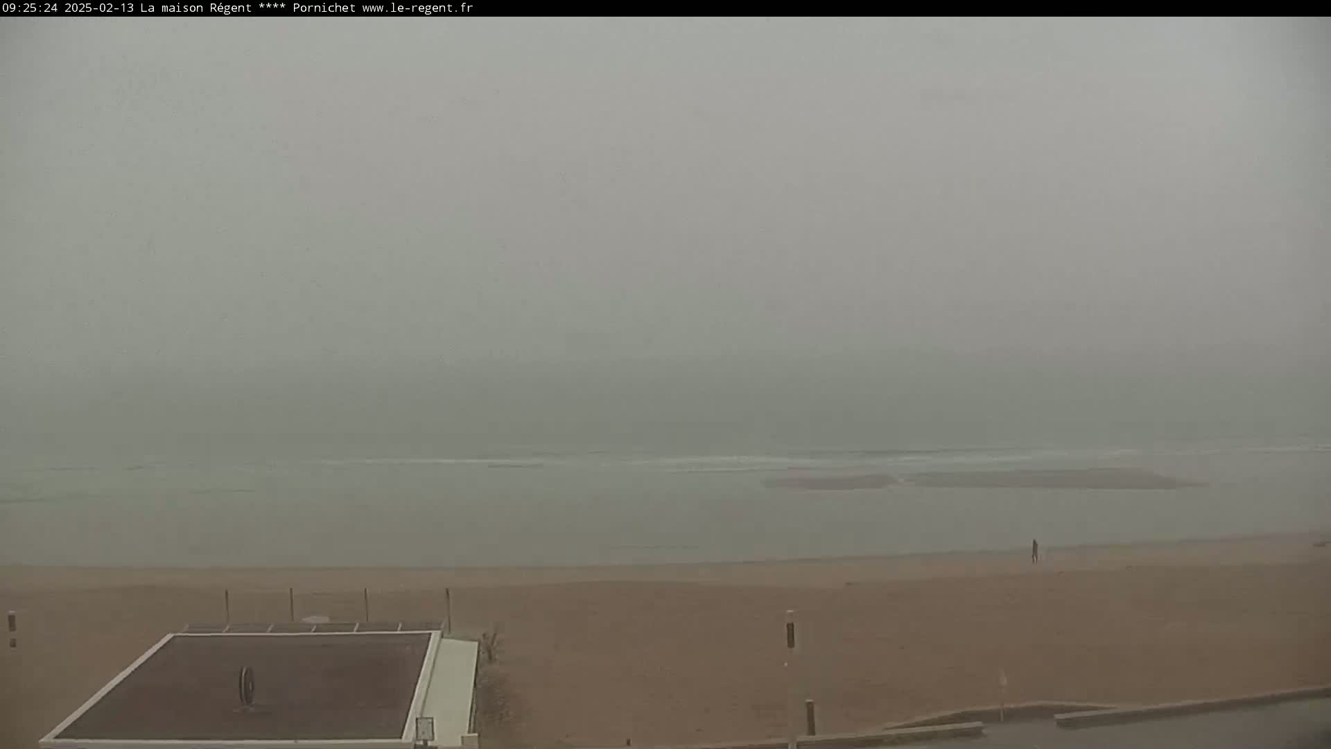 A hazy, overcast day shows a beach with calm waves, a lone person walking on the sand, and a dark-topped structure near the water's edge.
