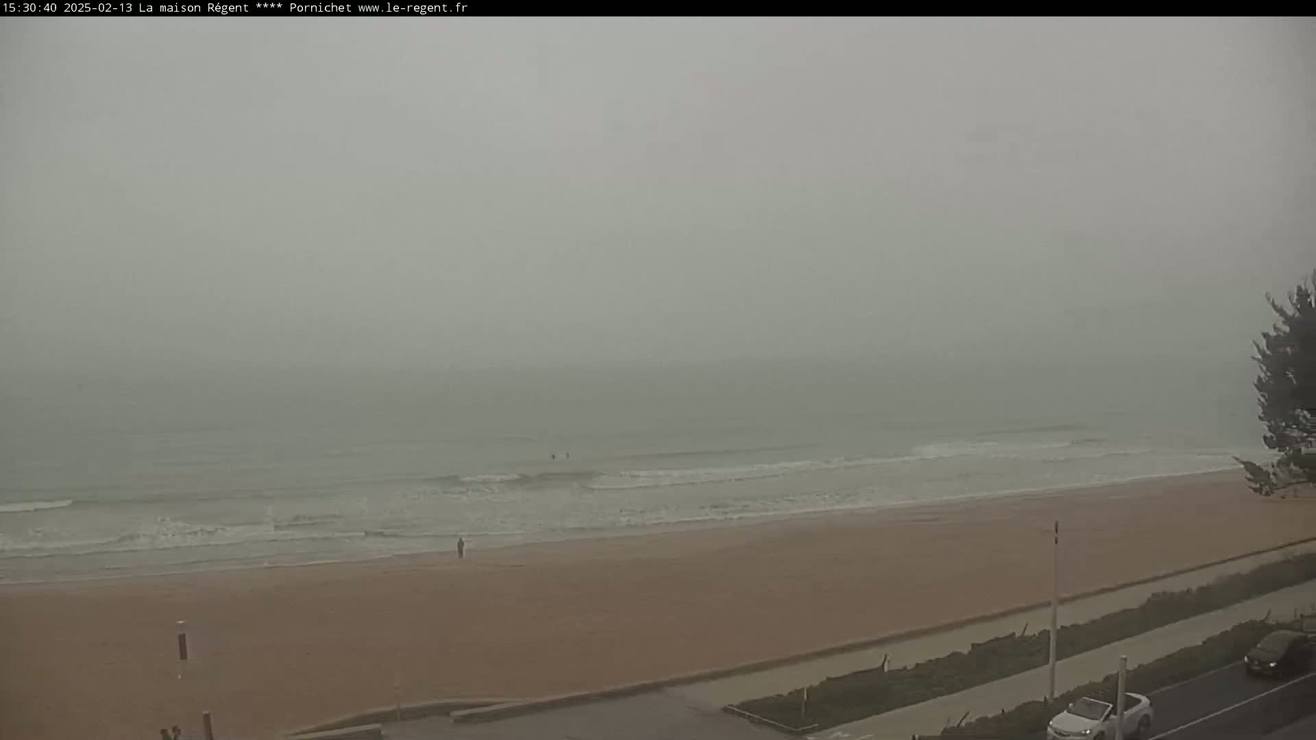 A hazy, overcast day reveals a sandy beach with small waves lapping the shore, a few people visible in the distance, and a road with cars parked along the side.