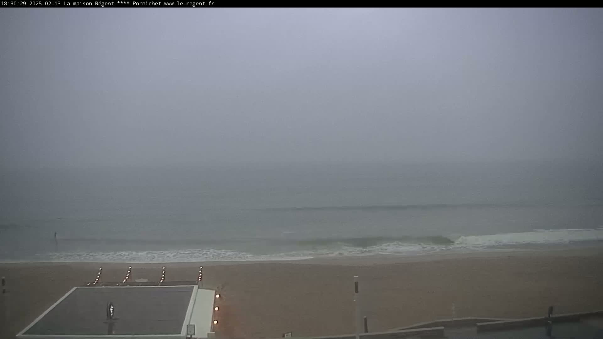 A foggy beach scene shows a calm ocean with small waves lapping at a sandy shore, where a flat, dark surface is partially visible near some lights.