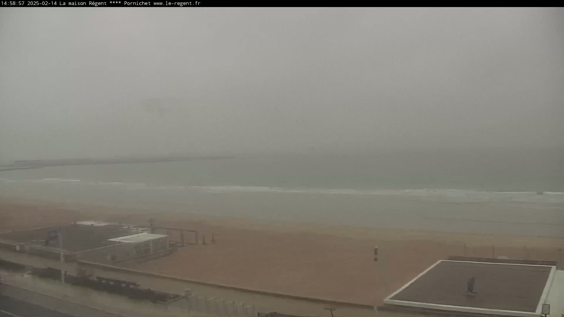 A hazy, overcast day shows a beach with a calm ocean and a few small structures along the shoreline.