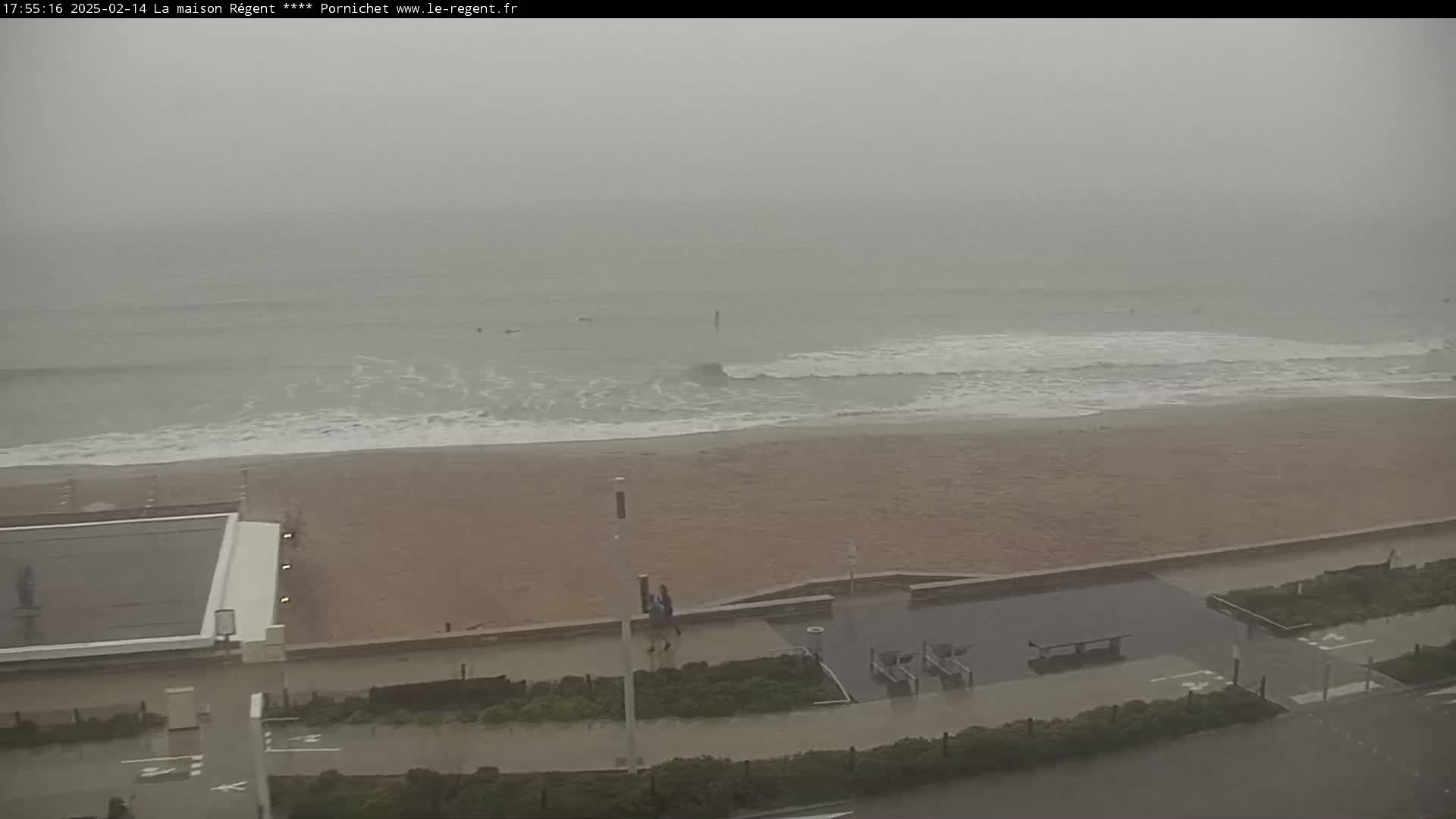 A foggy, rainy day at a beach shows waves lapping a sandy shore with a paved walkway and some people walking near a building.