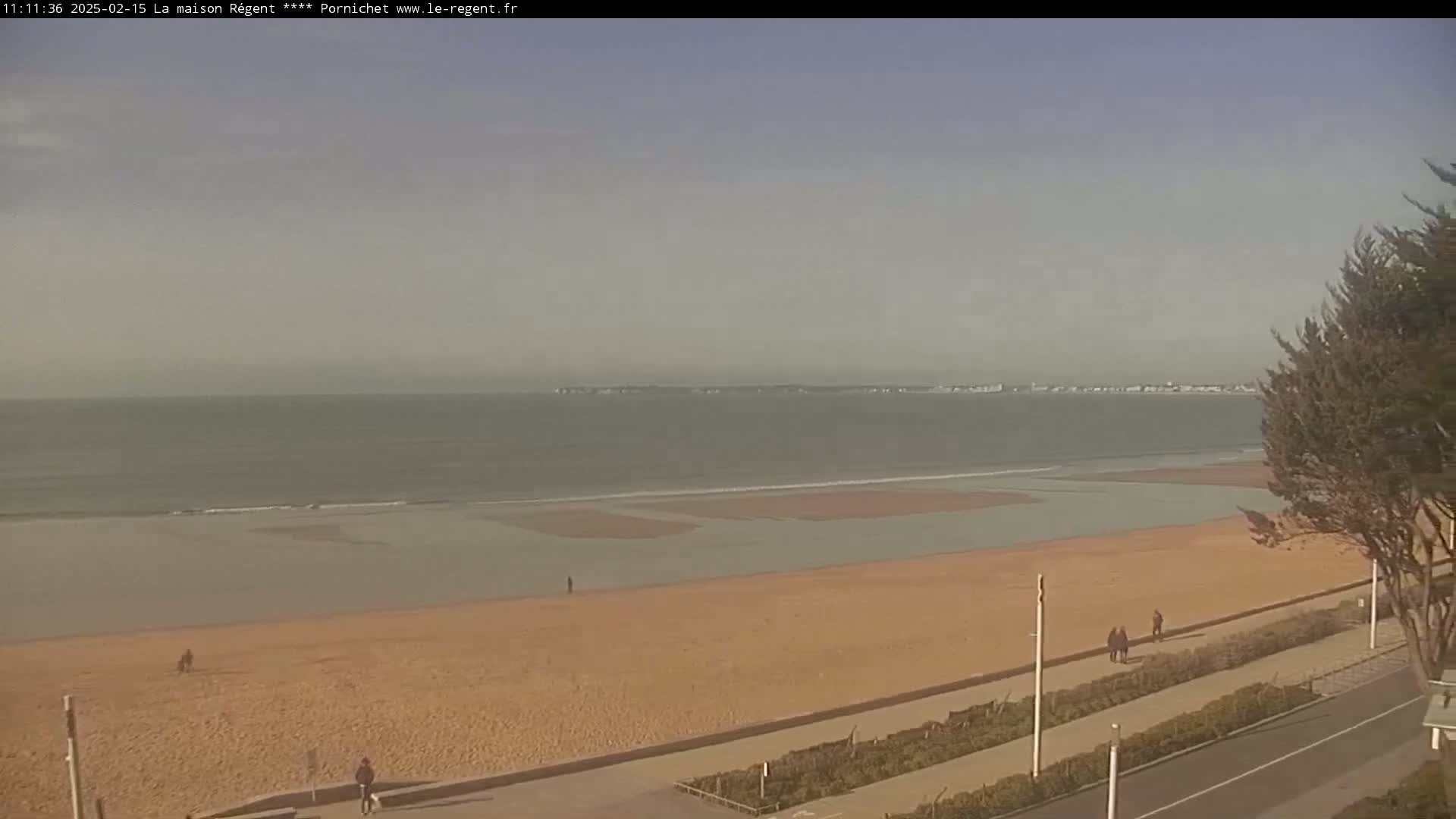 A mostly clear day reveals a sandy beach with a few people walking, leading to a calm ocean with a distant shoreline under a hazy sky.