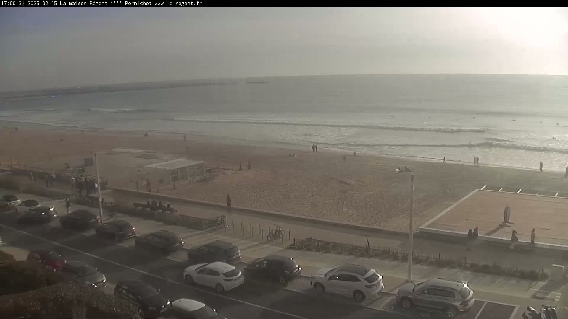 A sandy beach with a few people on it is bordered by a paved walkway with parked cars, all under a hazy, overcast sky.